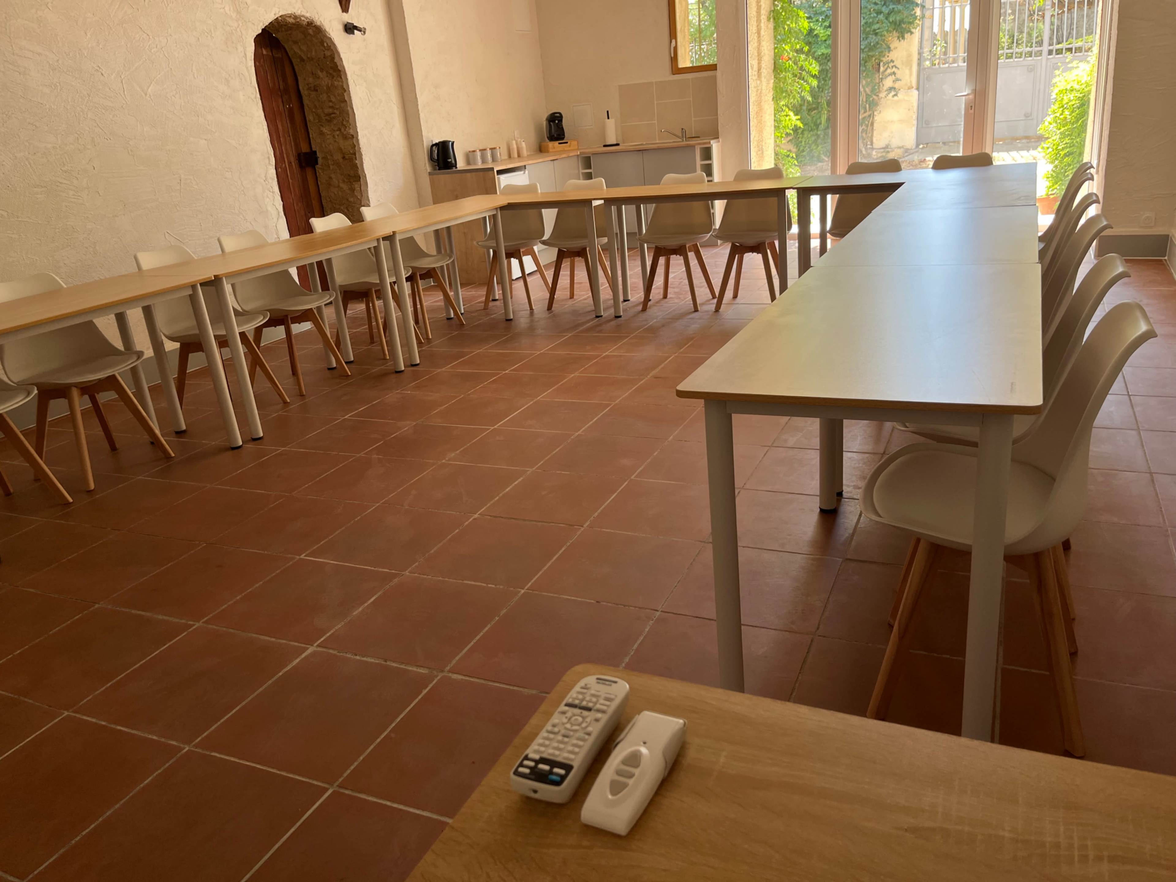 The image shows a spacious, sunlit room with multiple tables arranged in a U-shape and white chairs, featuring a remote control and a table organizer in the foreground.