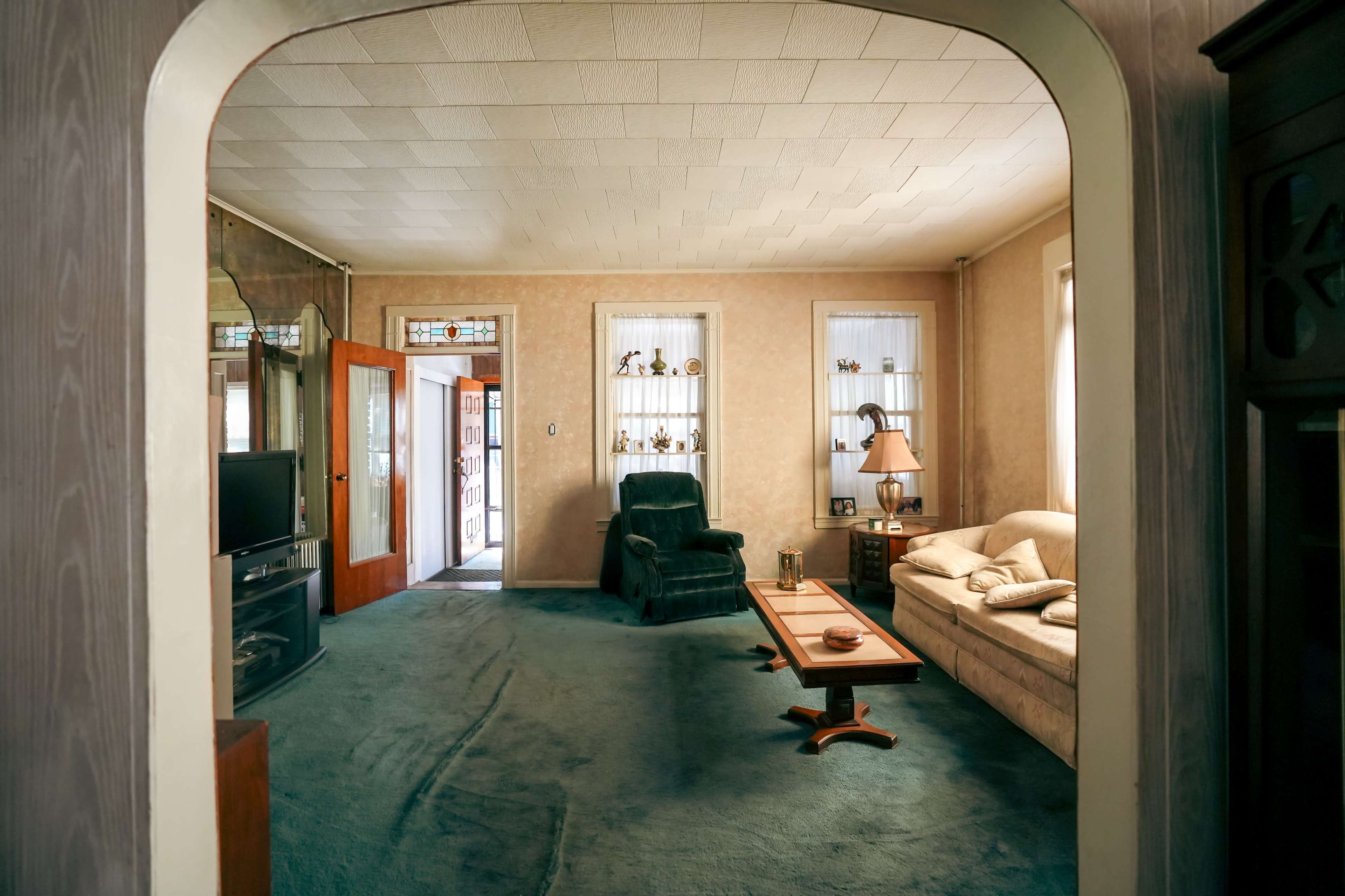 A living room features a blue carpet, a green armchair, a beige sofa, and shelves with decorative items.