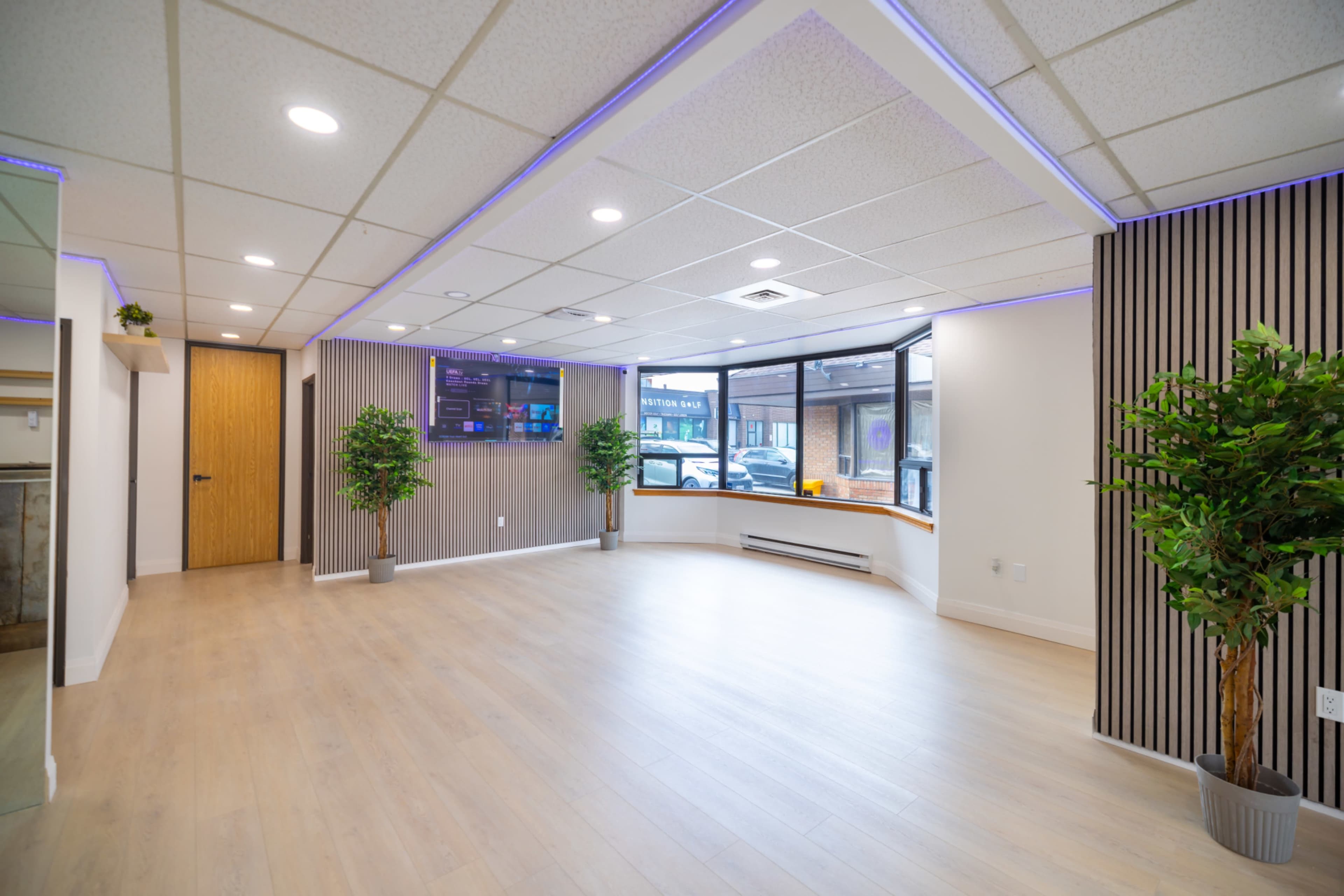 The image shows a spacious interior with wood flooring, large windows, and minimalist decor, including two potted plants and a wall-mounted screen.