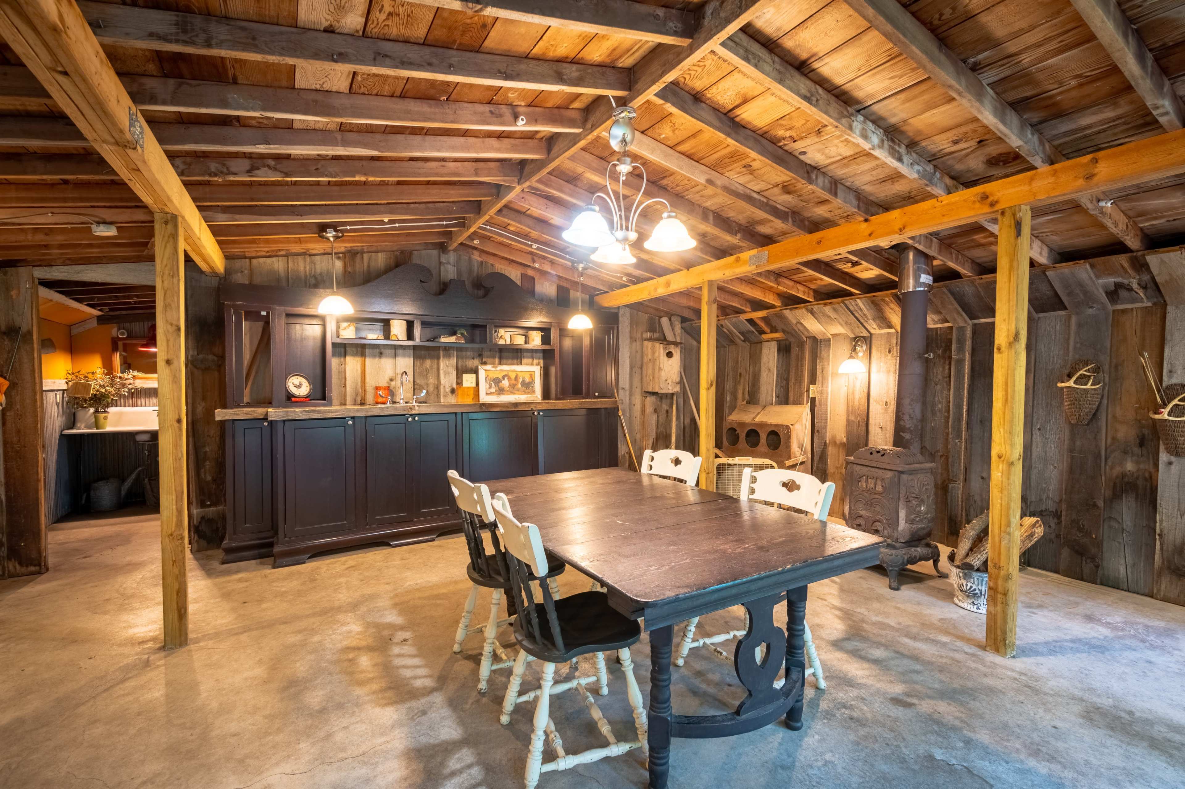 The interior of a rustic cabin features a wooden table with white chairs, a dark wooden bar unit, and a stove against the wall beneath a wooden ceiling.