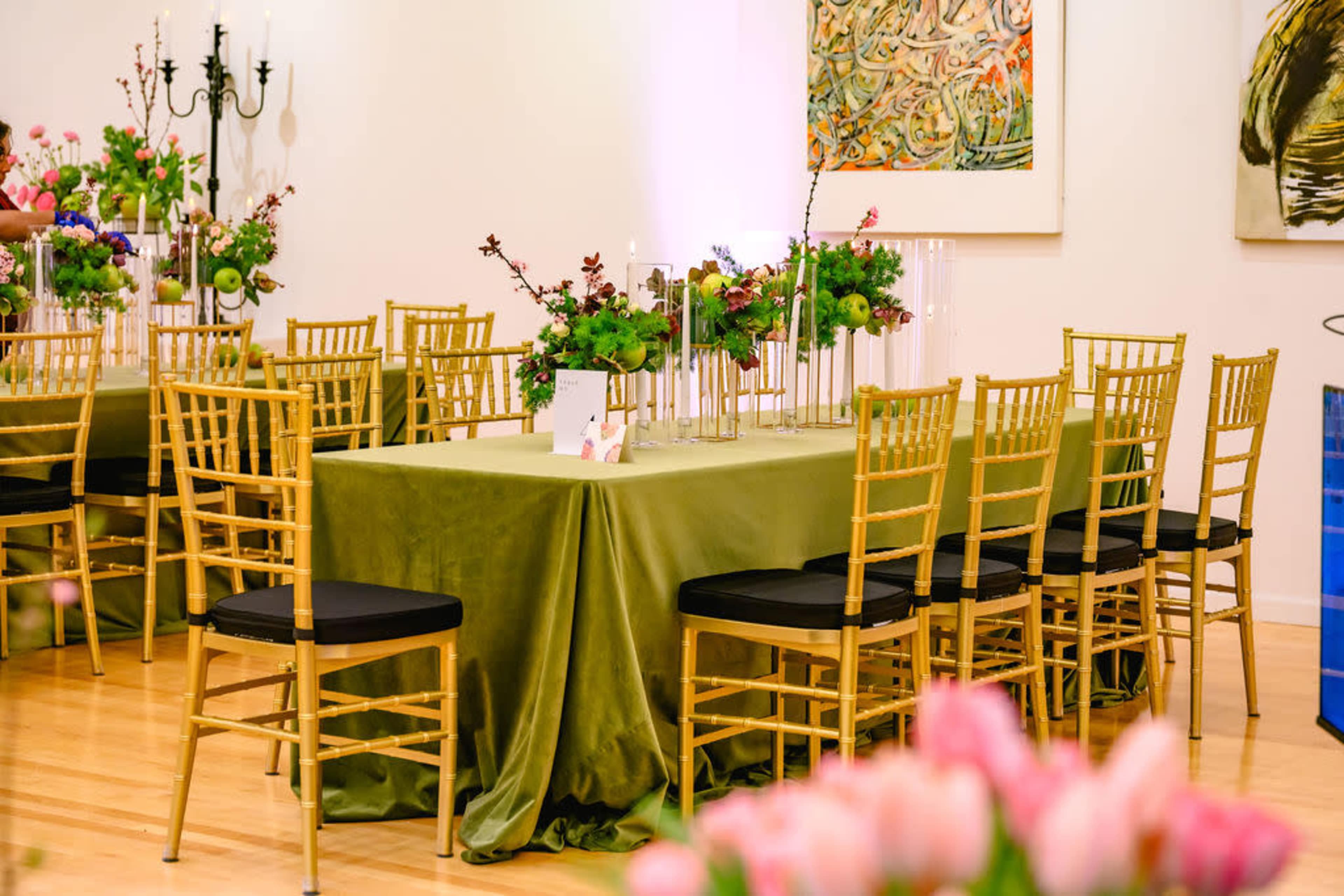 A dining table with a green tablecloth is set in a well-decorated room, surrounded by gold chairs and adorned with floral centerpieces.