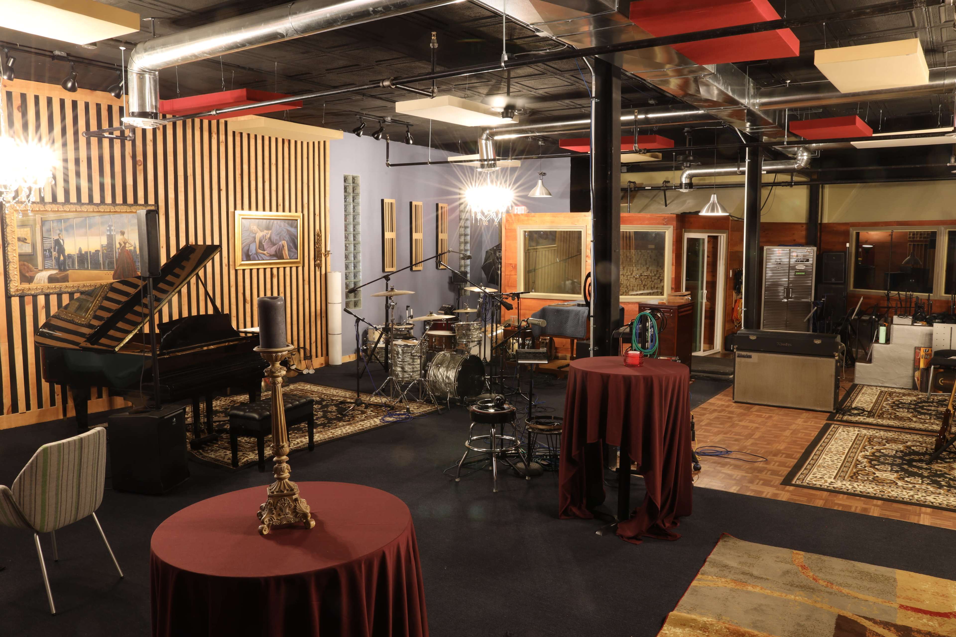 A music studio featuring a grand piano, a drum set, and several tables, with a cozy seating area and kitchen in the background.