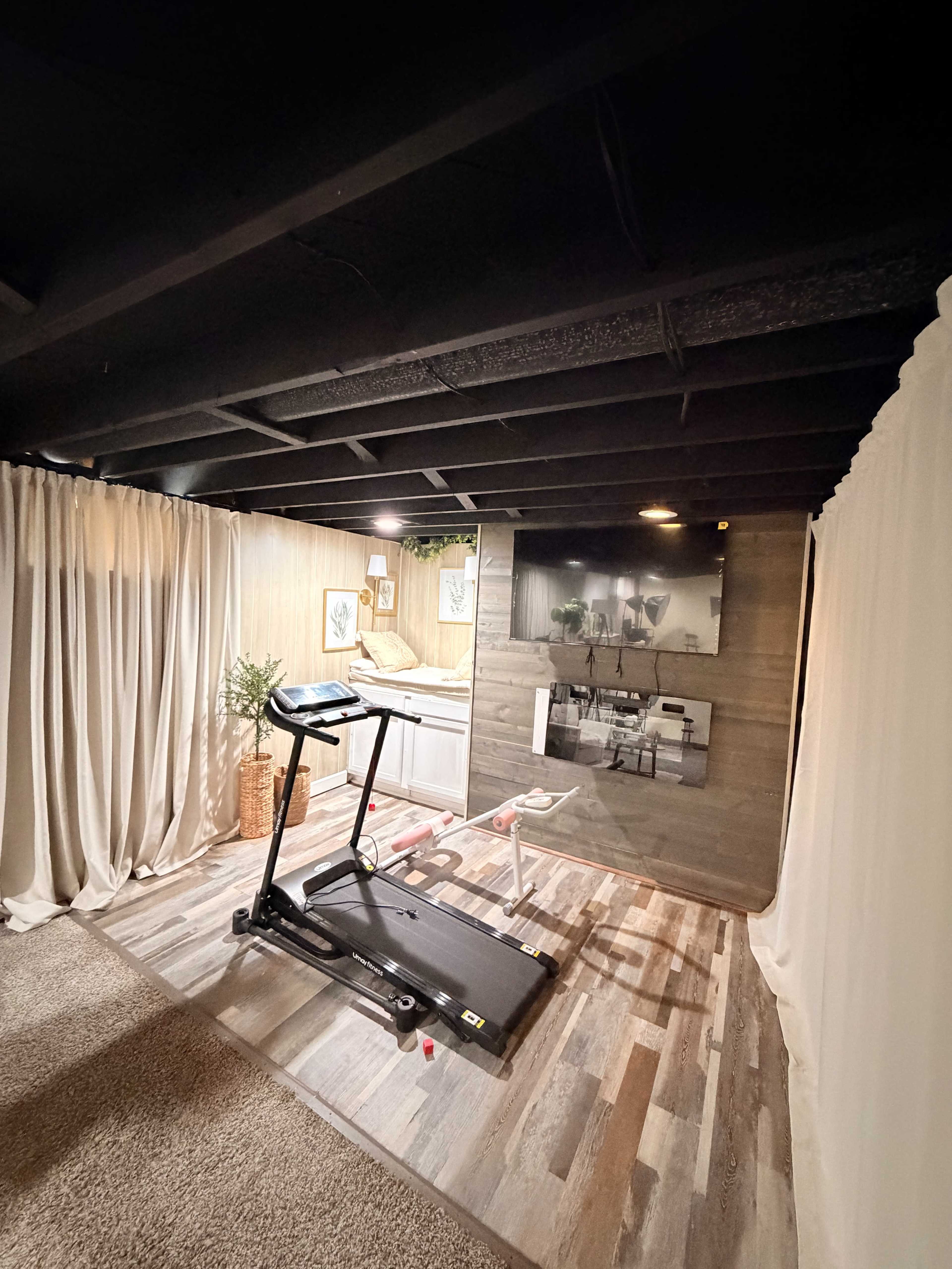 The image shows a home gym area with a treadmill, a muted wooden wall, and a television mounted above it, separated by curtains.
