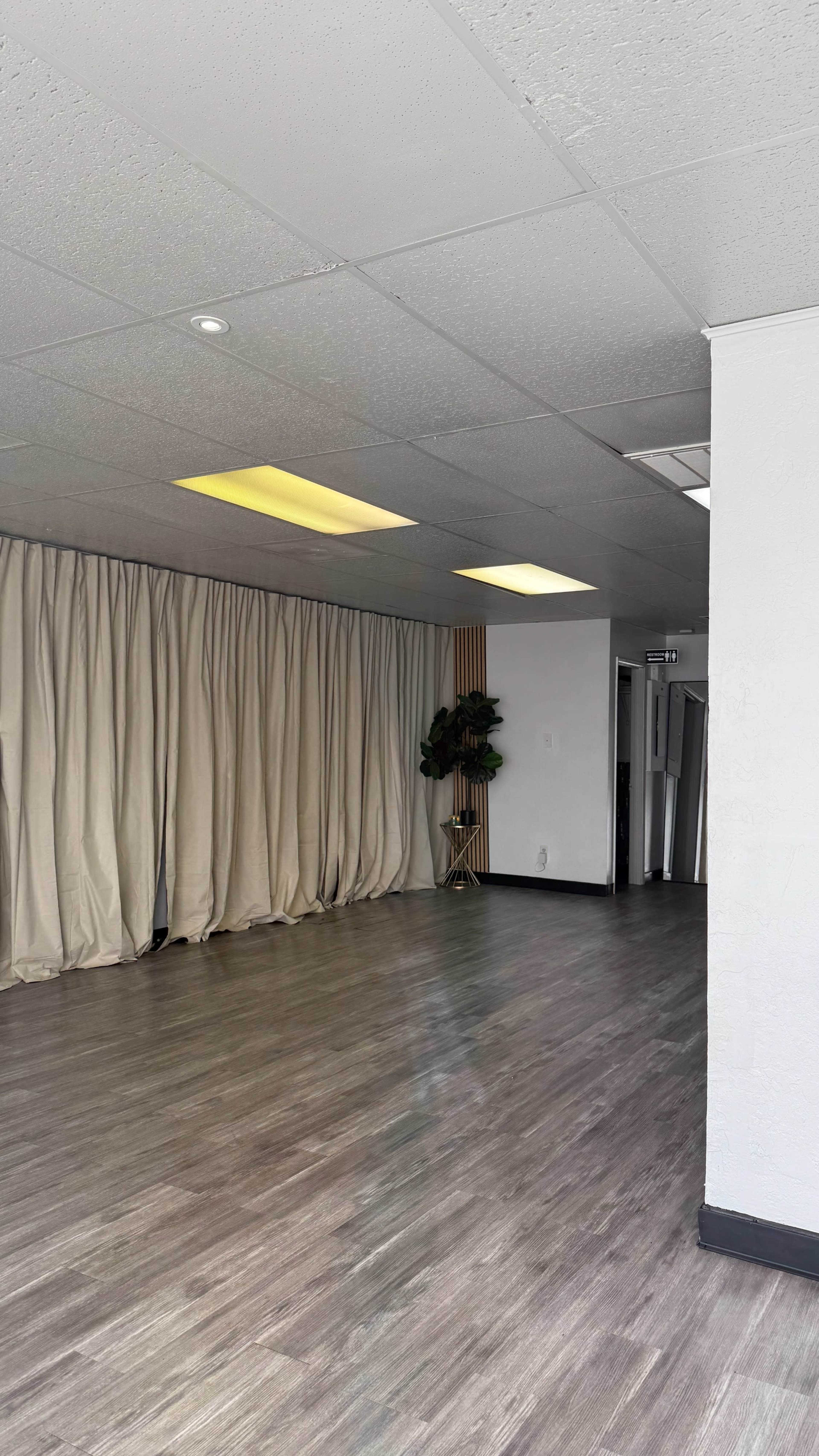 The image shows a large, empty indoor space with wooden flooring, beige curtains along one wall, and a plant in a corner.