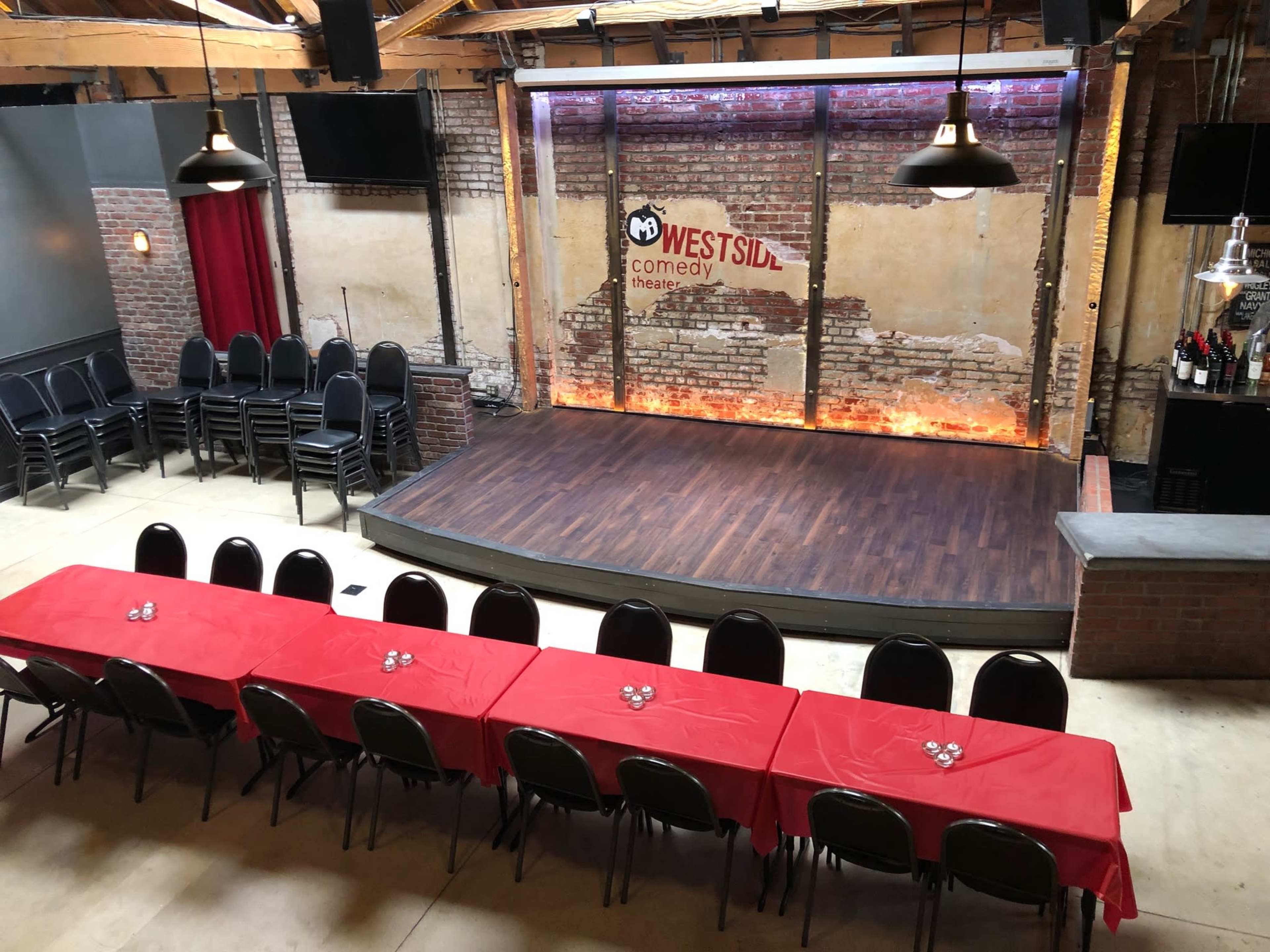 A comedy theater setup featuring a stage with a brick backdrop, surround lighting, and rows of black chairs facing a long red table arranged for an audience.