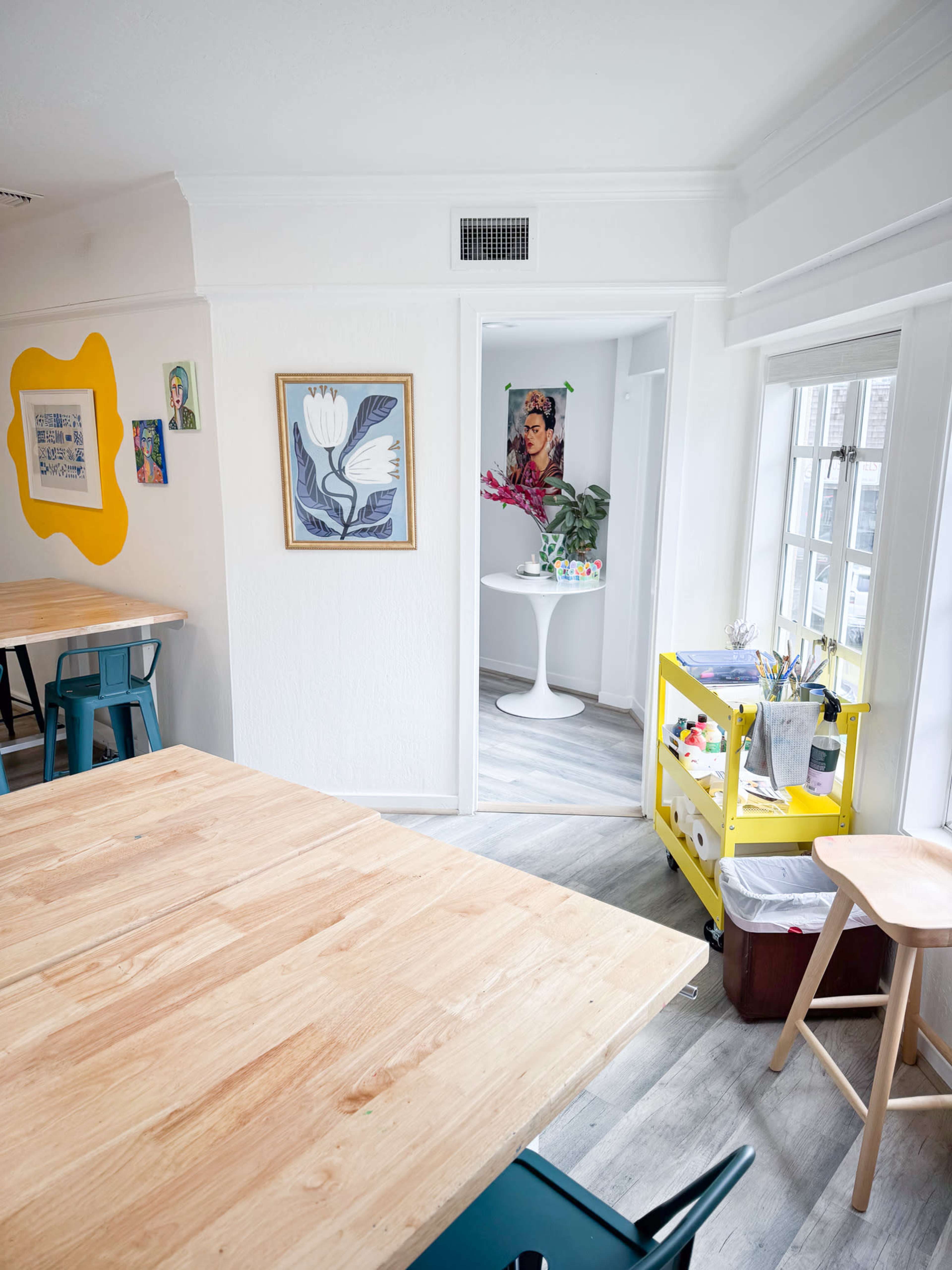 A bright, modern workspace with wooden tables, a vibrant yellow wall art piece, and a small table displaying flowers and art supplies.
