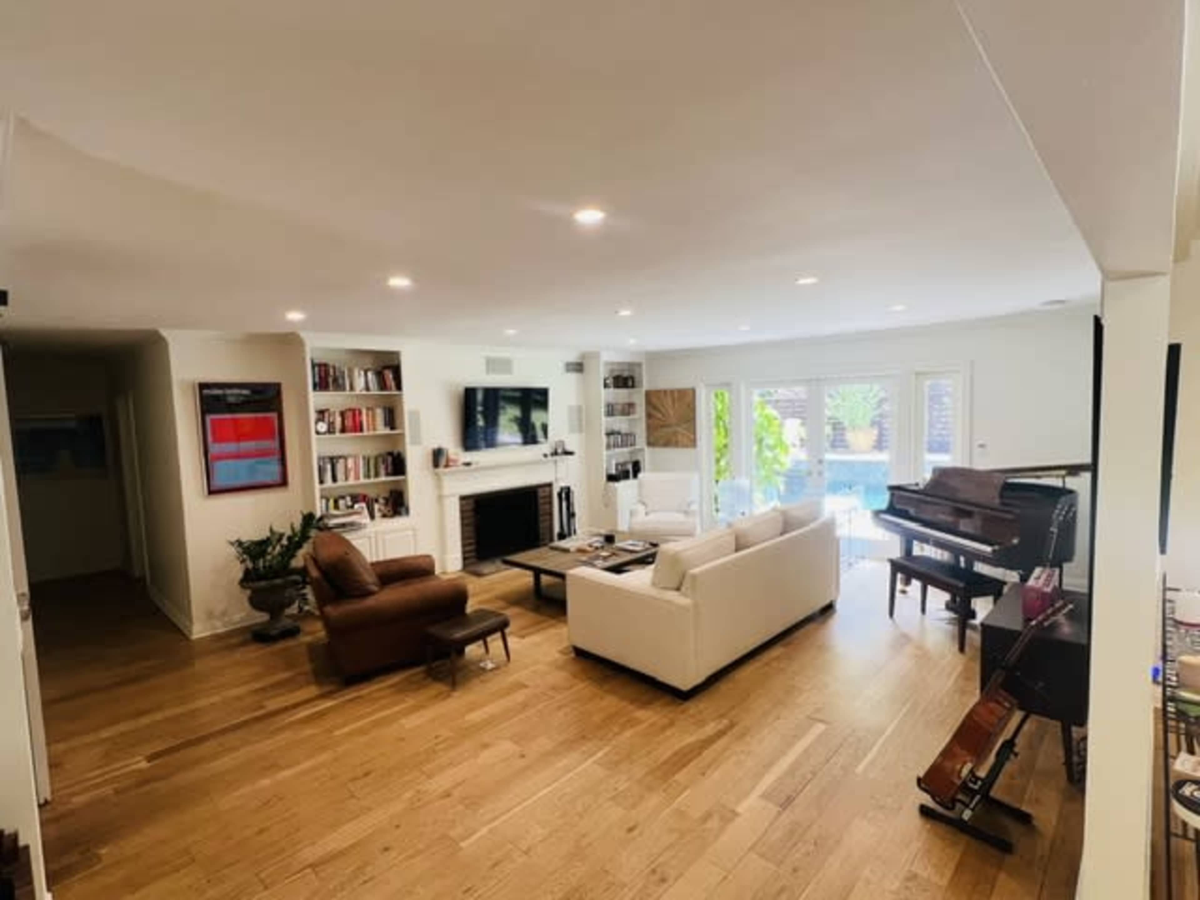 A spacious living room features a white sofa, a brown armchair, a piano, and large windows overlooking a pool area.
