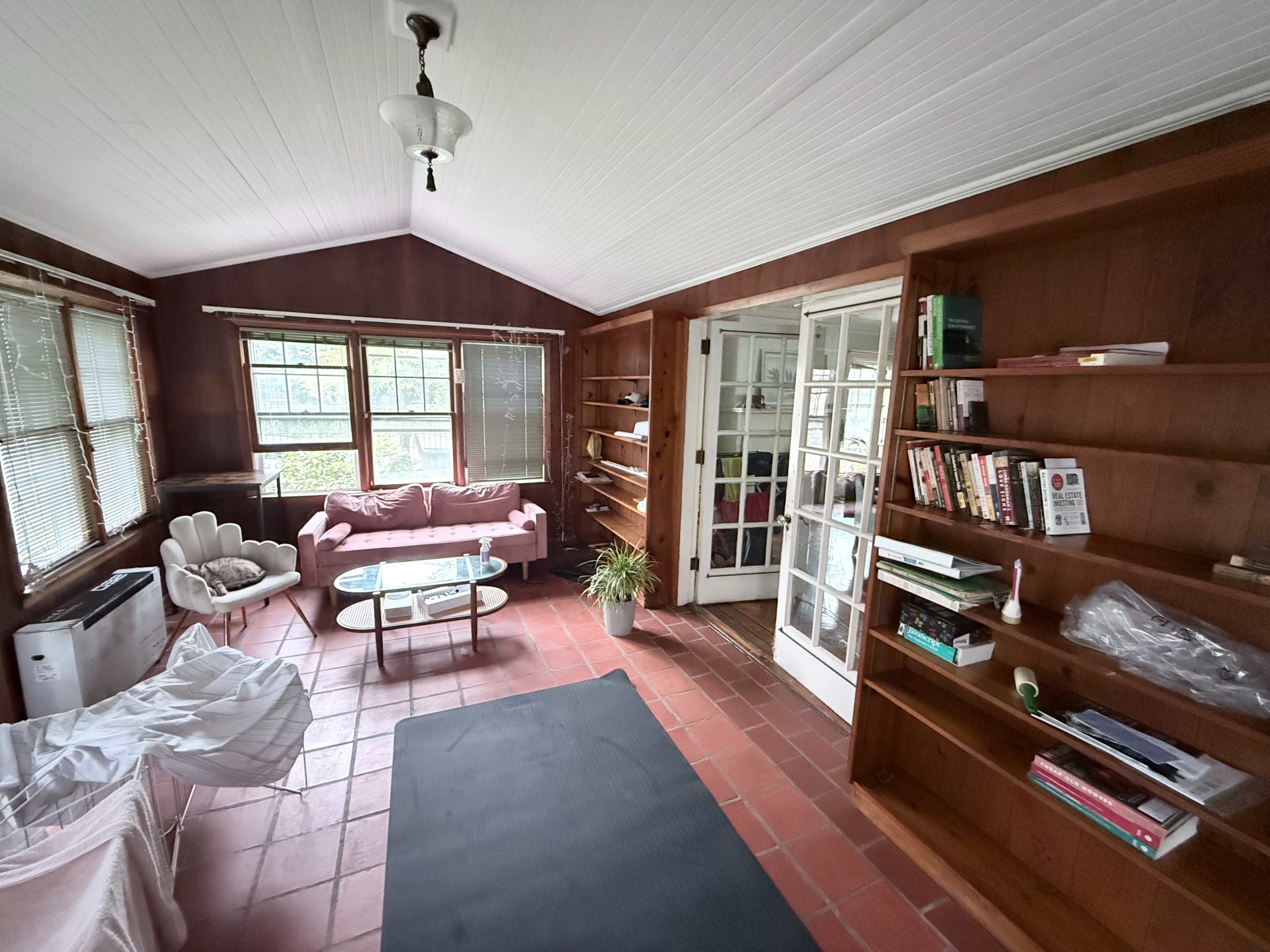 The image shows a cozy living space with wooden walls, a pink couch, a glass coffee table, and shelves filled with books.