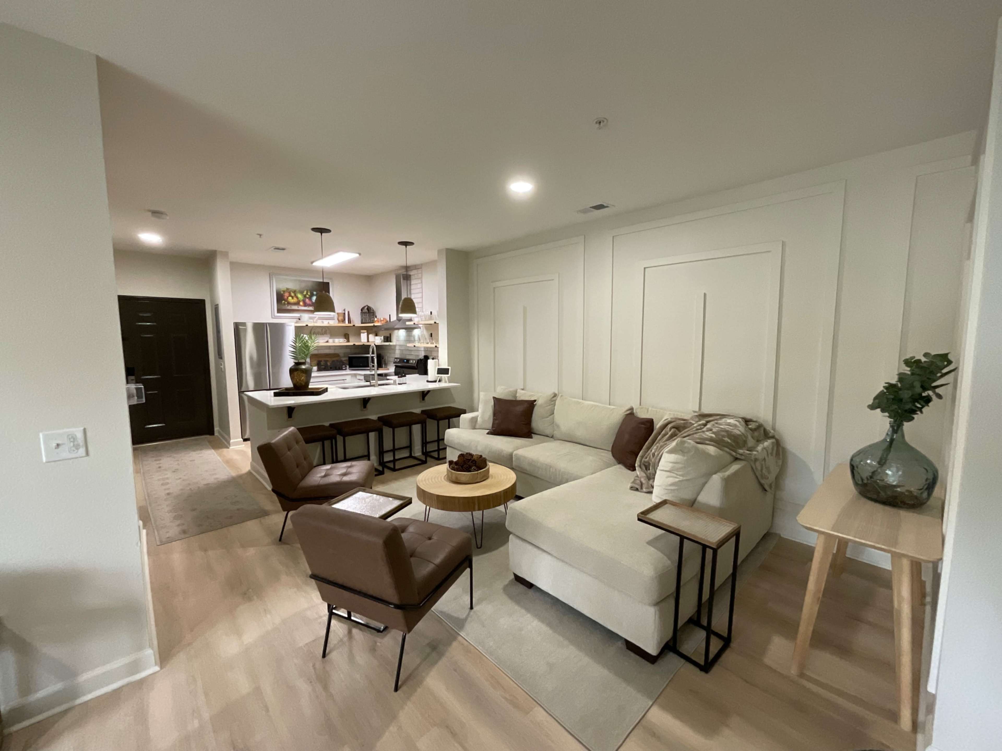 A modern living room with a sectional sofa, two brown chairs, a round coffee table, and a view of a kitchen with bar stools in the background.