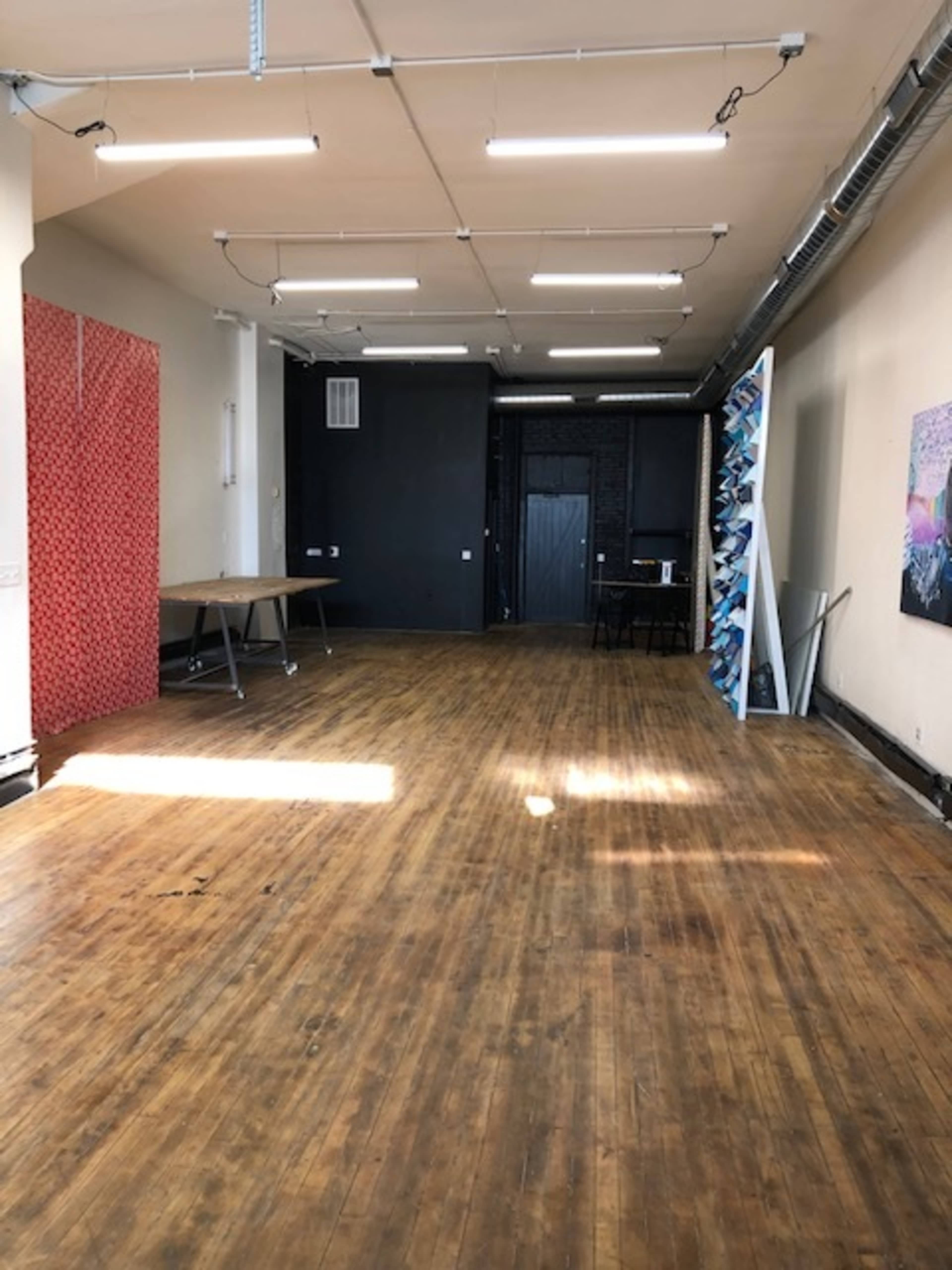 An empty room with wooden floors, a large black wall, a red patterned divider, and several tables and artworks arranged along the walls.