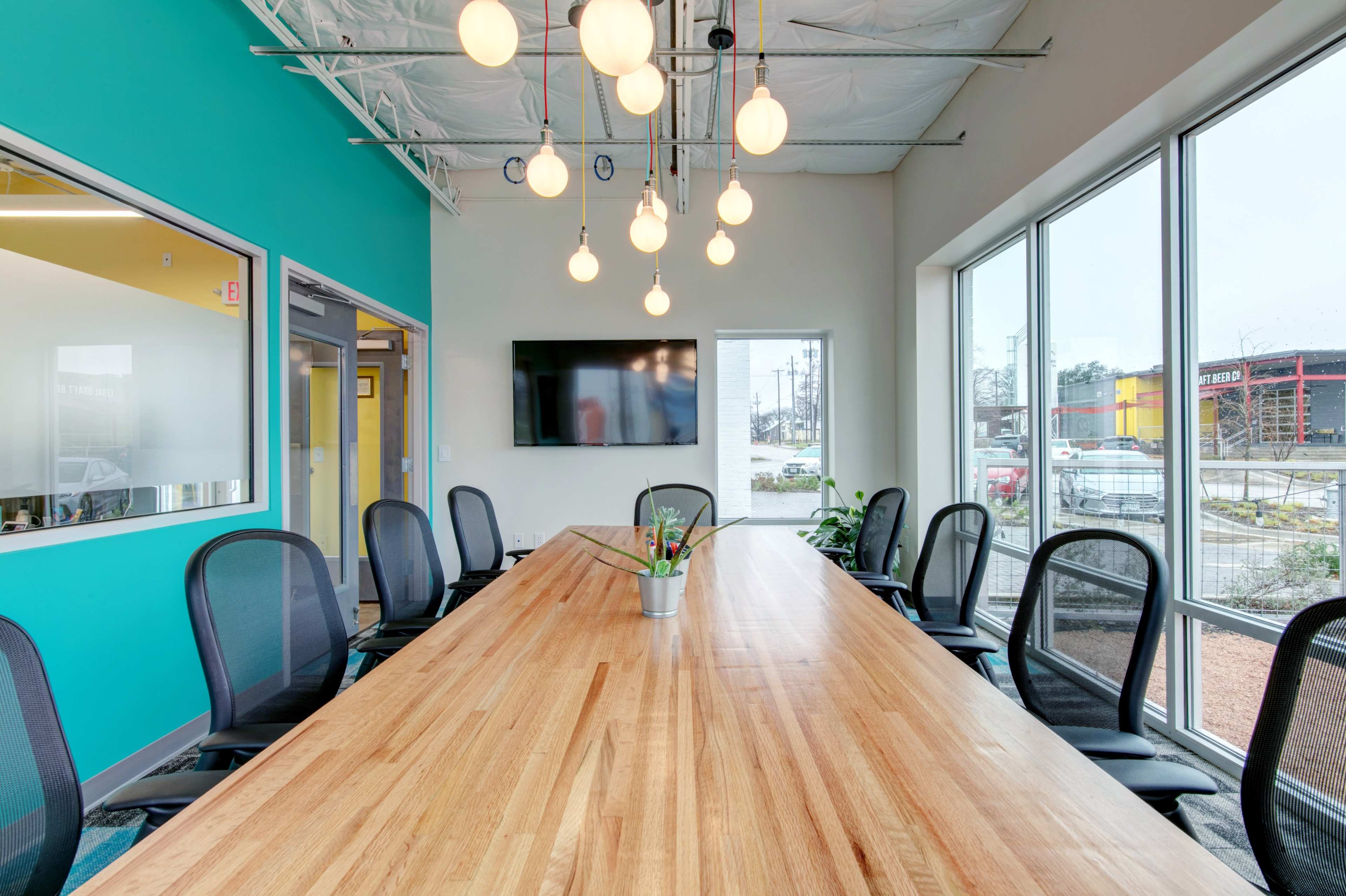 A large conference room features a wooden table surrounded by black office chairs, with a turquoise wall and modern lighting fixtures overhead.