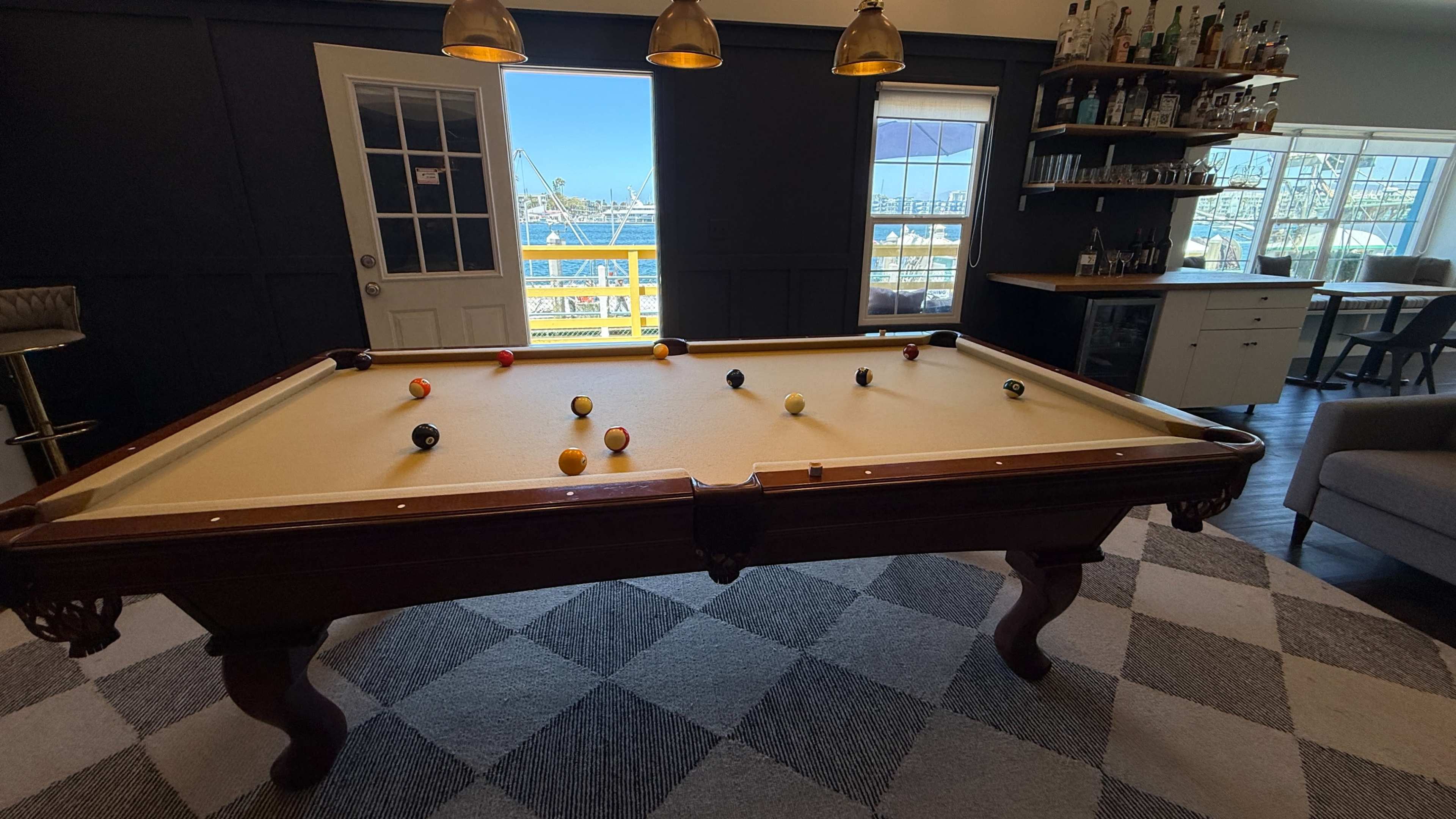 A billiard table with balls arranged on it is positioned in a room overlooking a marina through large windows.