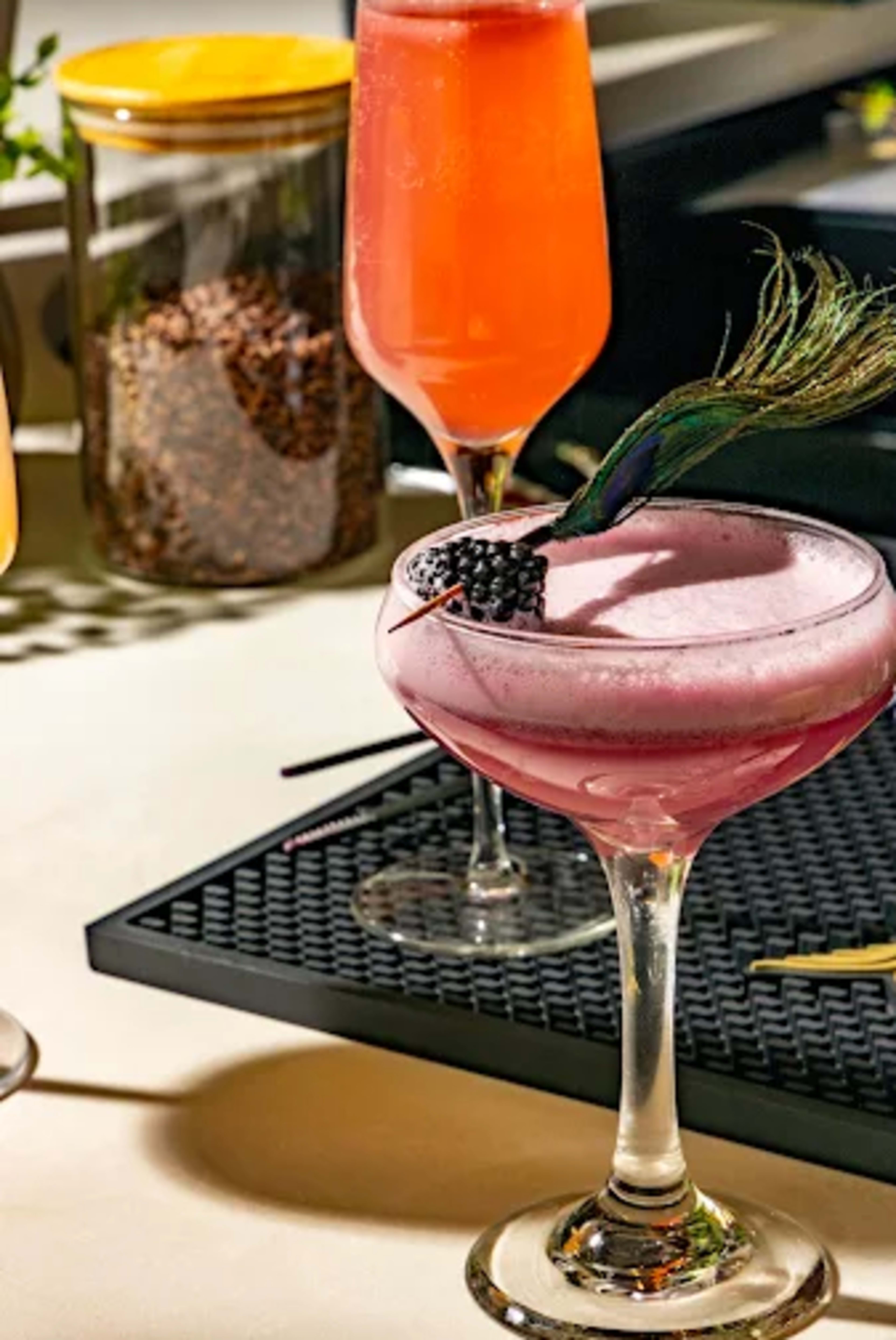 The image features three colorful cocktails, each served in elegant glasses, with a garnish of fruit and a decorative peacock feather on one.