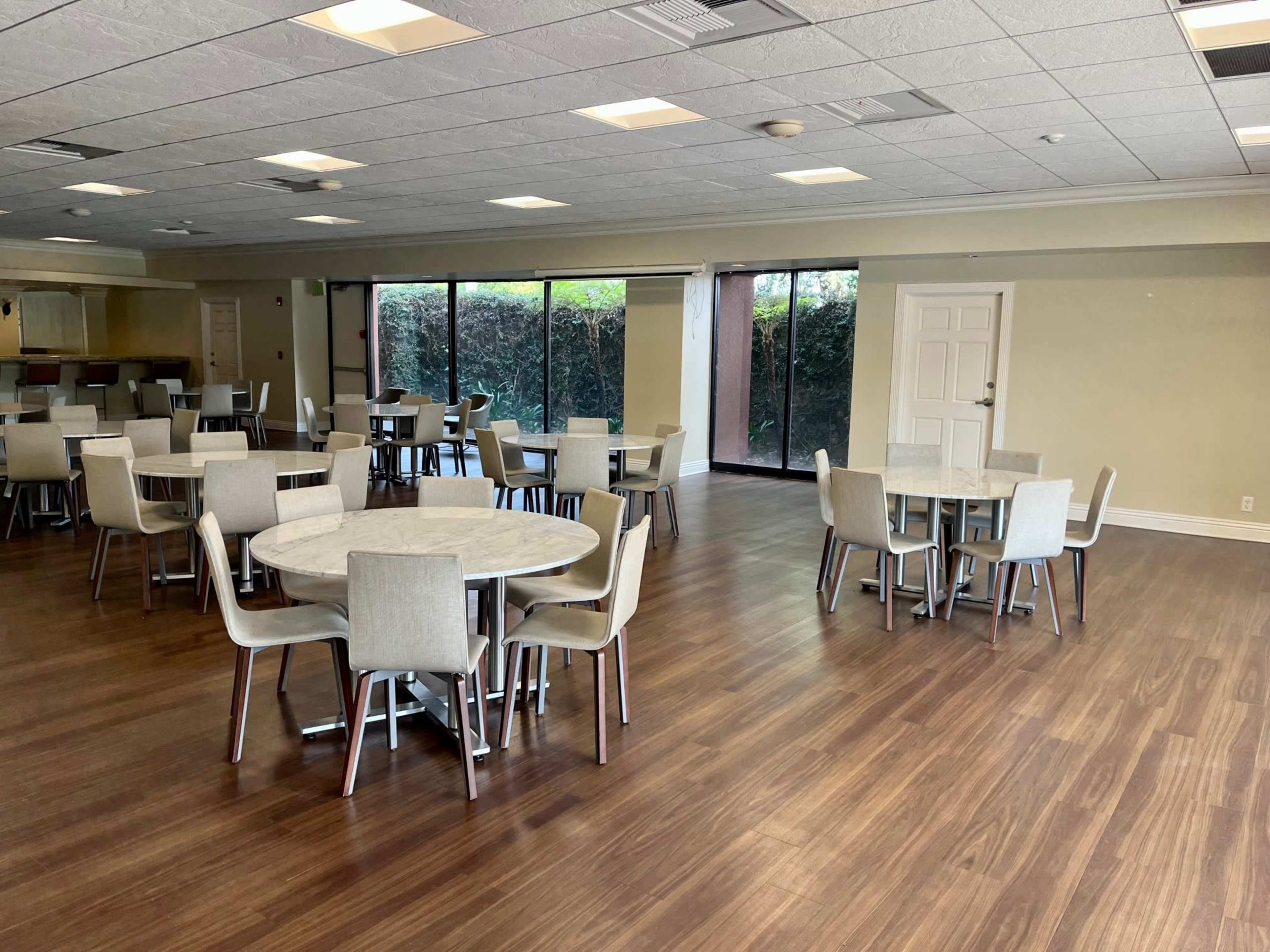A spacious dining area features circular tables and chairs arranged on a wooden floor, with large windows offering a view of greenery outside.