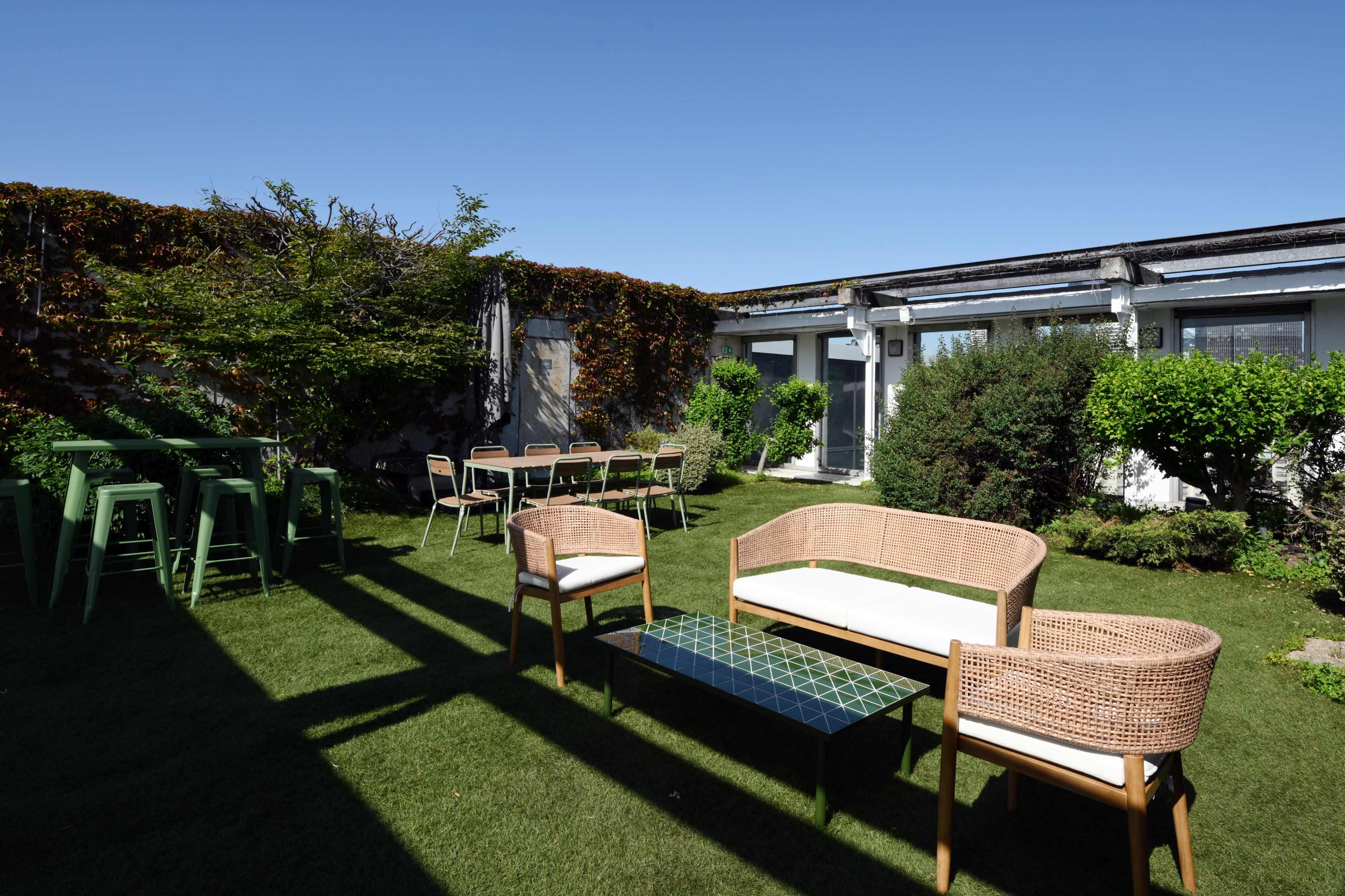 The image shows a landscaped outdoor area with seating arrangements, including a rattan sofa and chairs, a glass-top table, and a row of tables and chairs in the background.