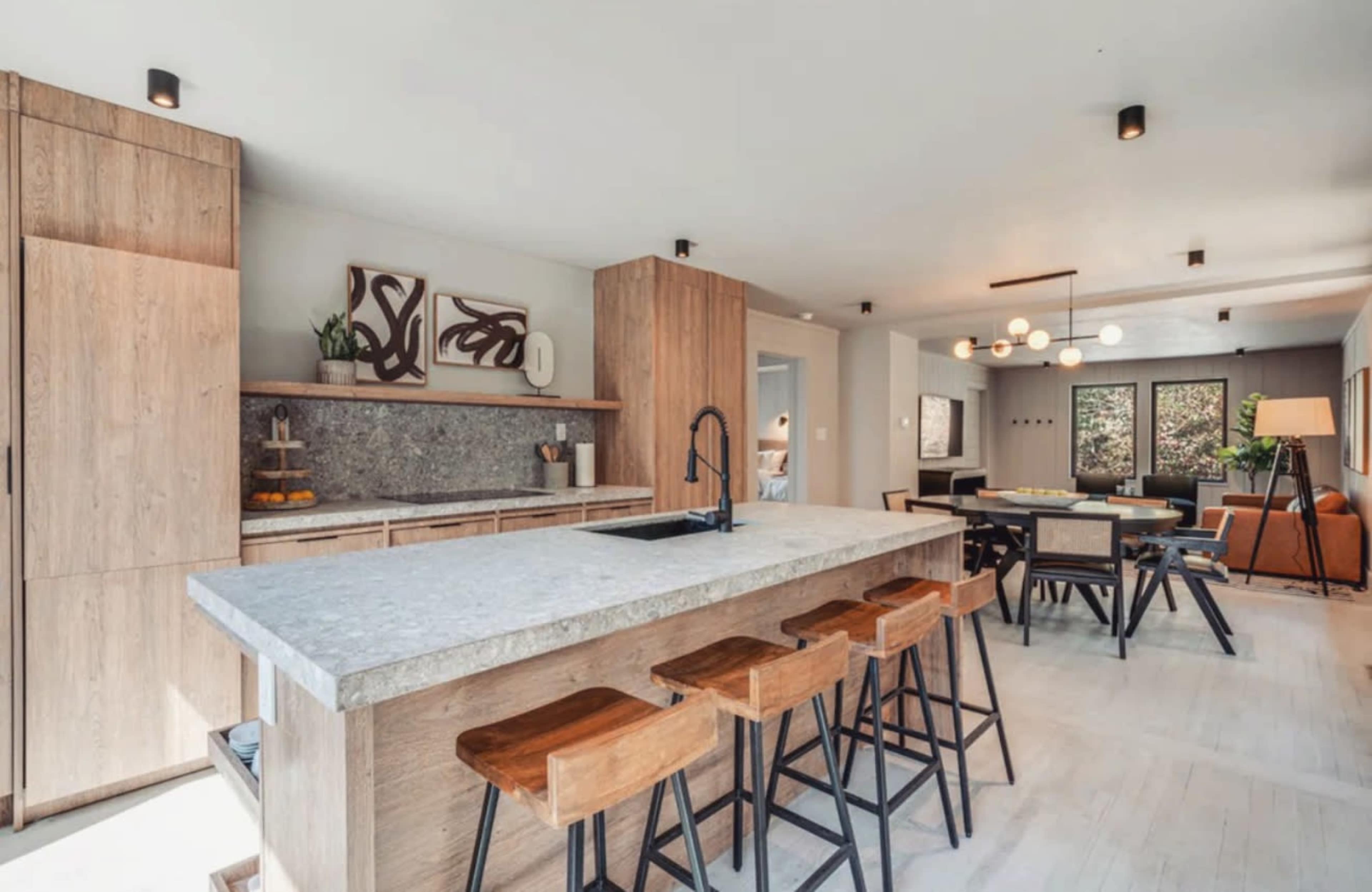 The image depicts a modern kitchen featuring a large island with gray countertops, wooden cabinetry, and a dining area with a table and chairs.