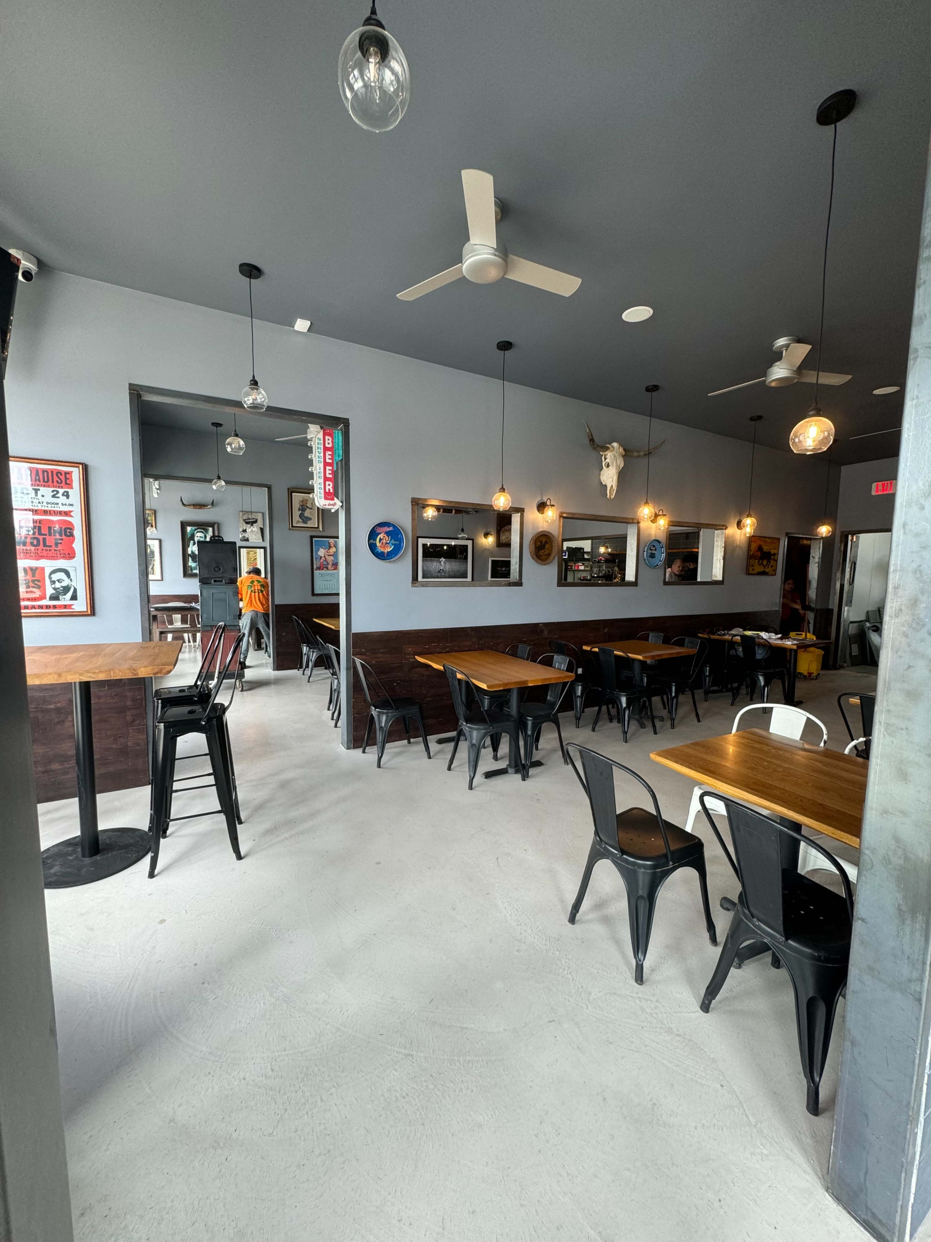 The interior of a restaurant features wooden tables, metal chairs, and ceiling fans, with a few decorative elements on the walls.