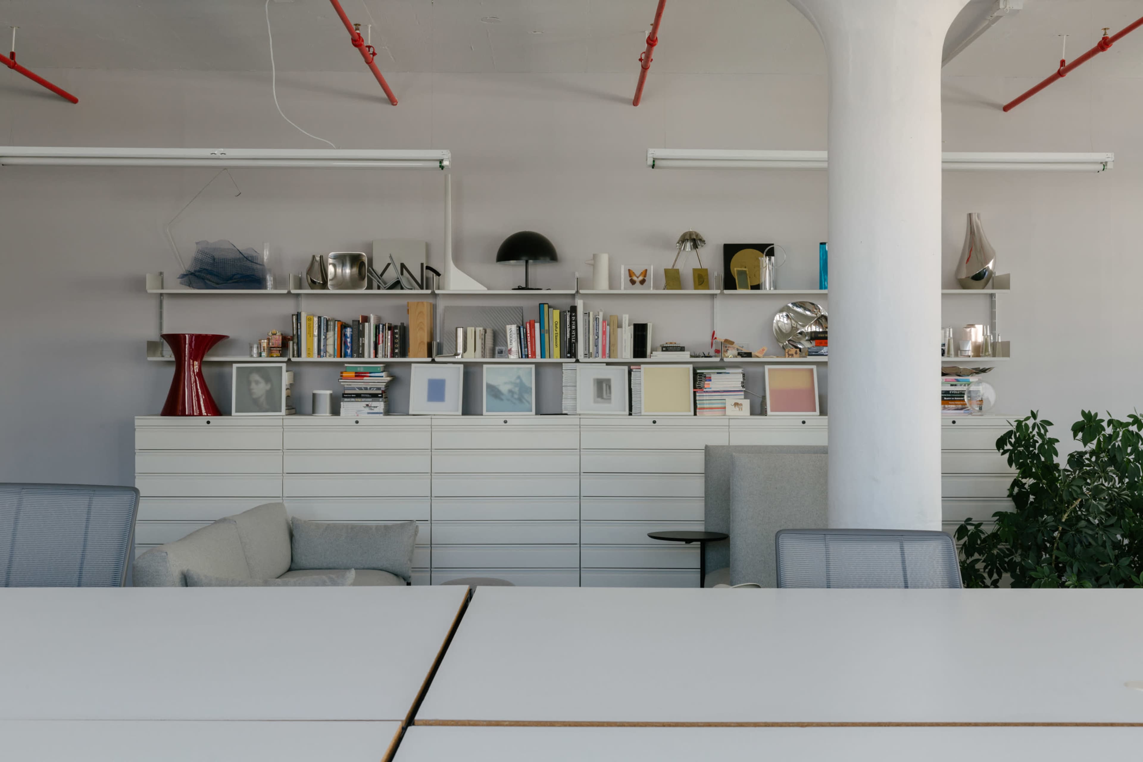 A modern office interior featuring a white shelving unit filled with books and decorative items, a gray armchair, and a table with visible surface lines.