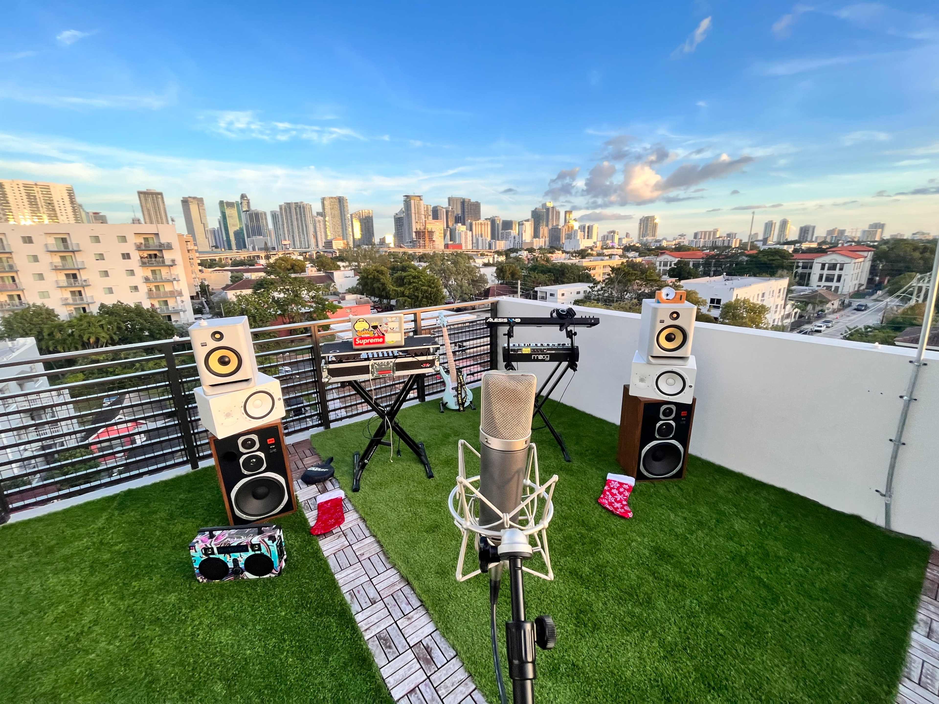 A rooftop scene with a microphone and various music equipment set up on artificial grass, overlooking a city skyline.
