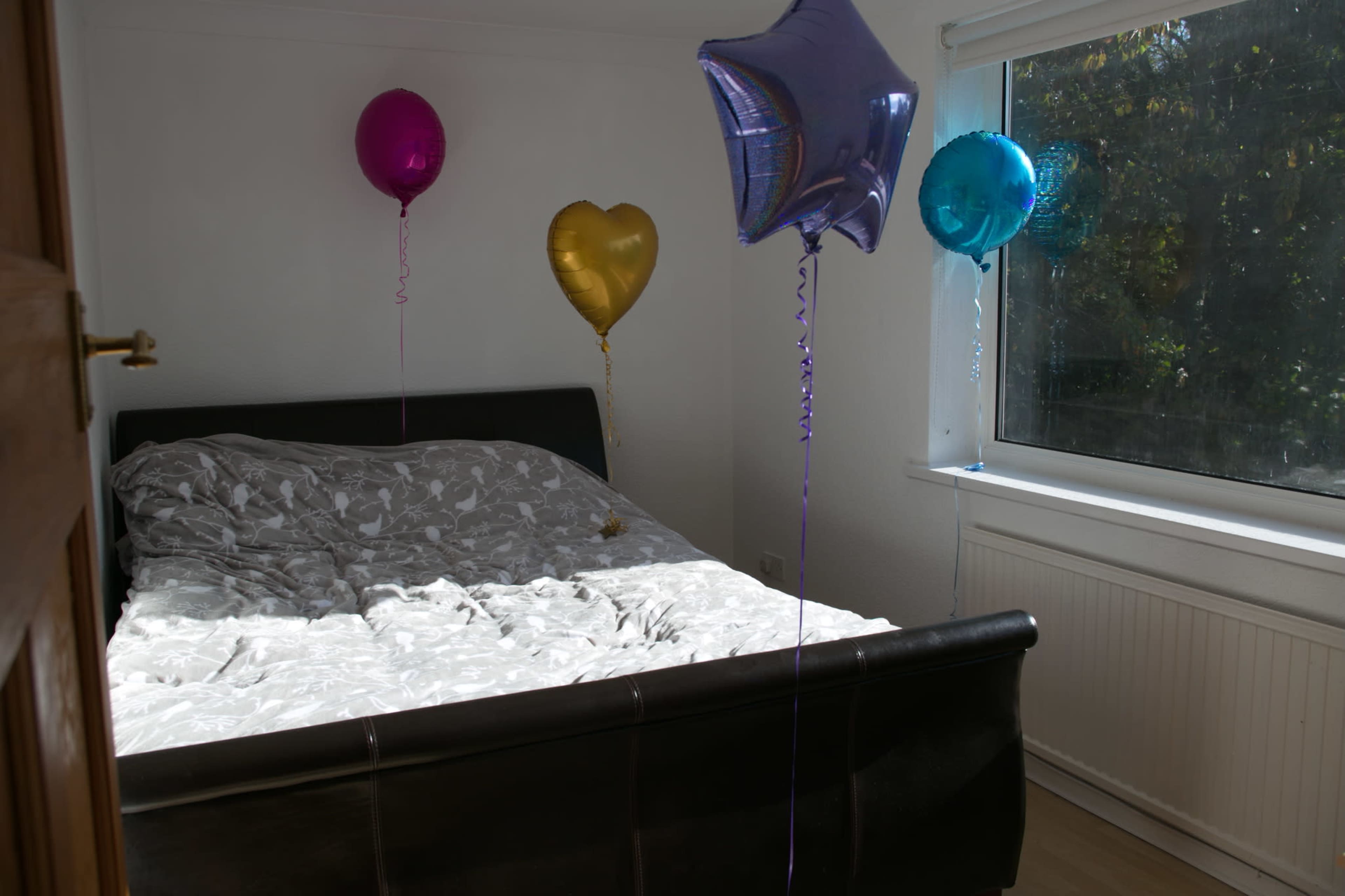 A bed with a patterned cover is positioned in a well-lit room, surrounded by colorful balloons of various shapes.