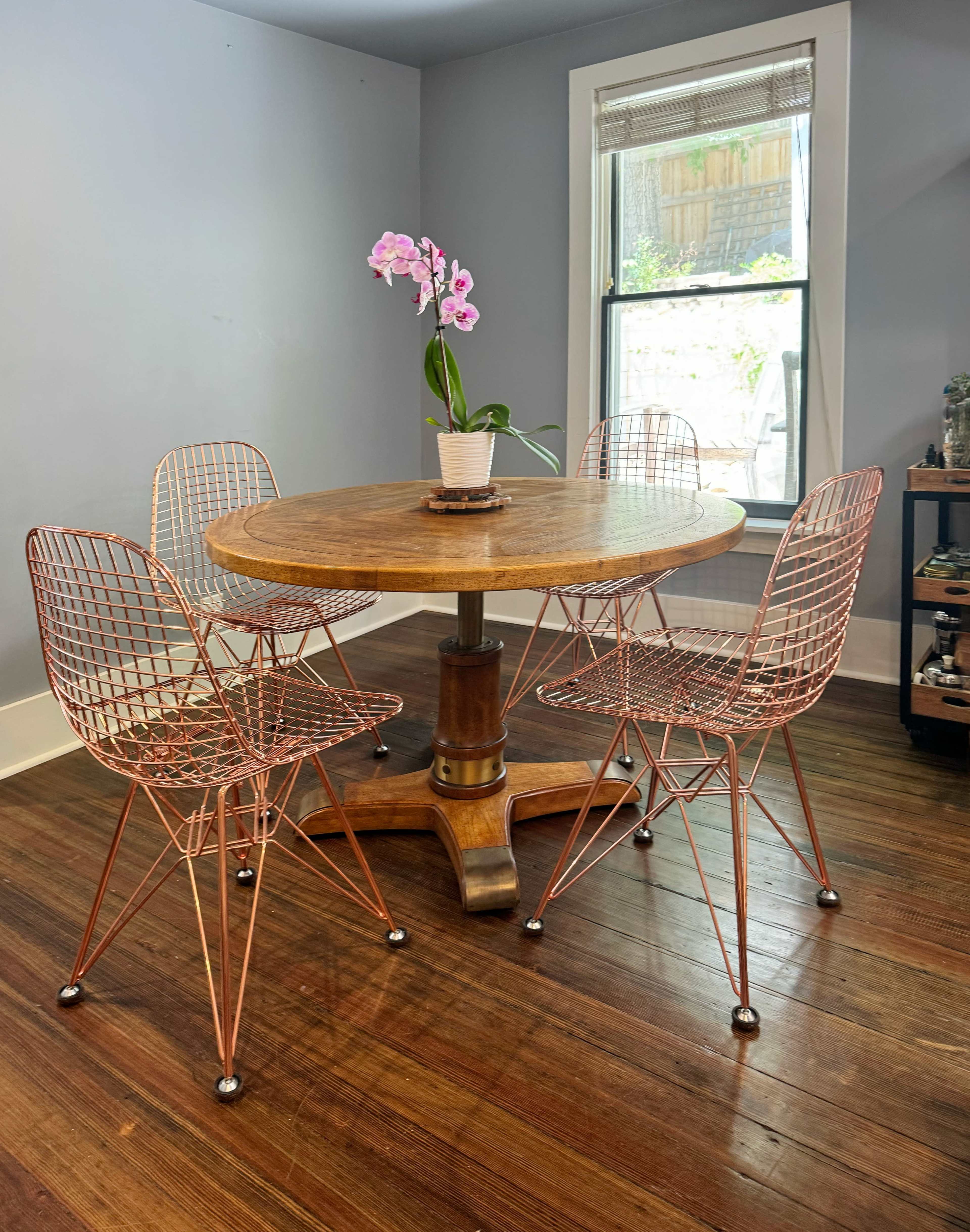 A round wooden table with a central pedestal is surrounded by four wire-frame chairs, and an orchid in a pot sits in the middle of the table.