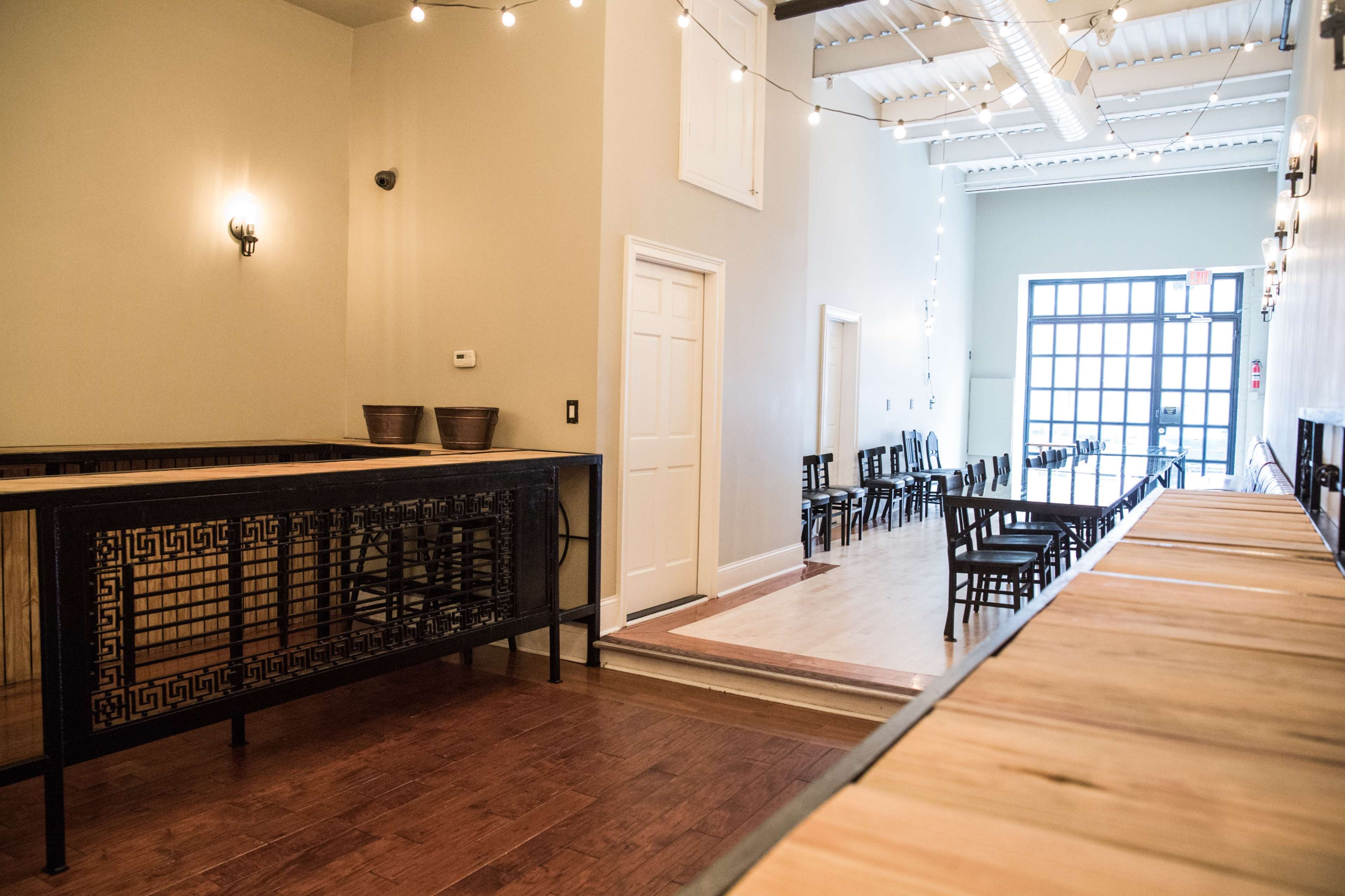 The interior of a spacious venue features a bar area on the left and rows of chairs arranged along the right side, illuminated by string lights above.