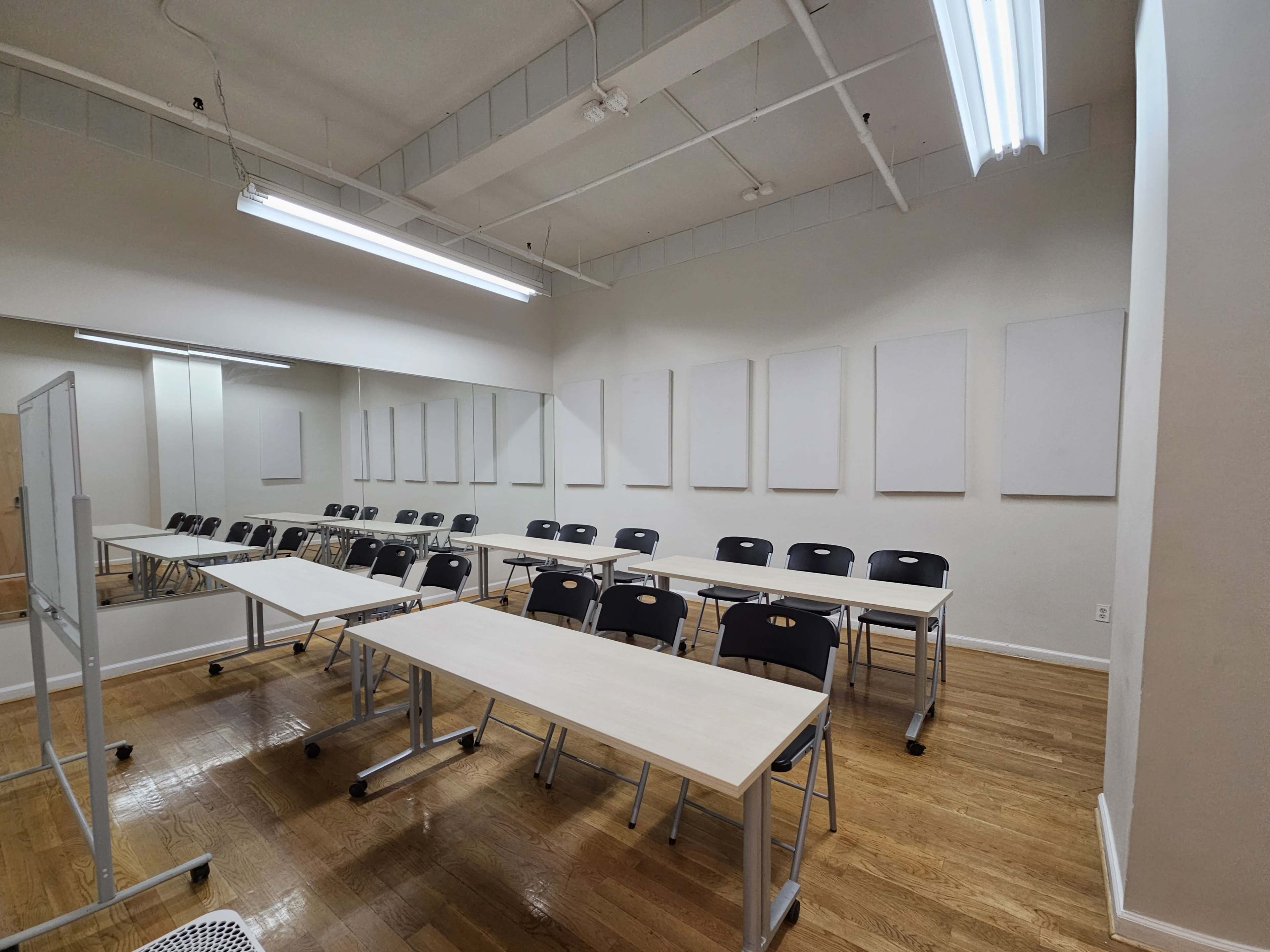 A sparsely furnished classroom with rows of tables and chairs, along with blank white wall panels and overhead lighting.