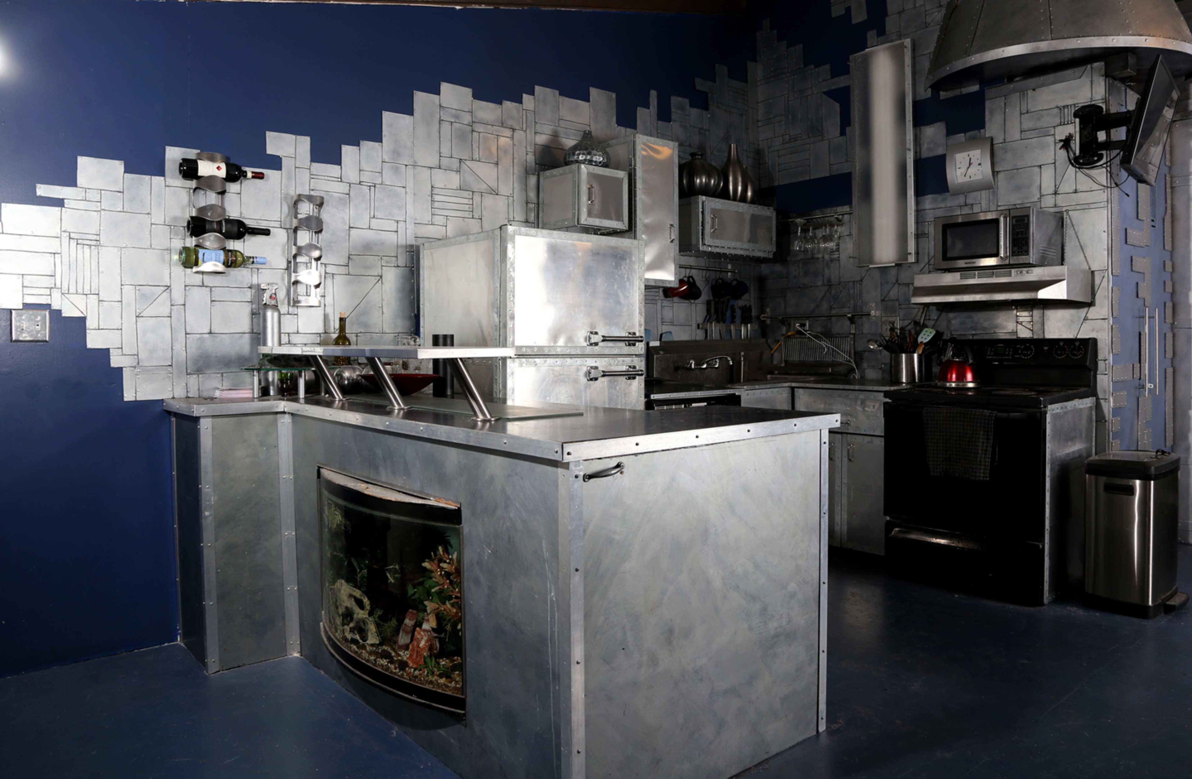 The kitchen features metallic surfaces, an aquarium integrated into the counter, and modern appliances against a geometric wall design.