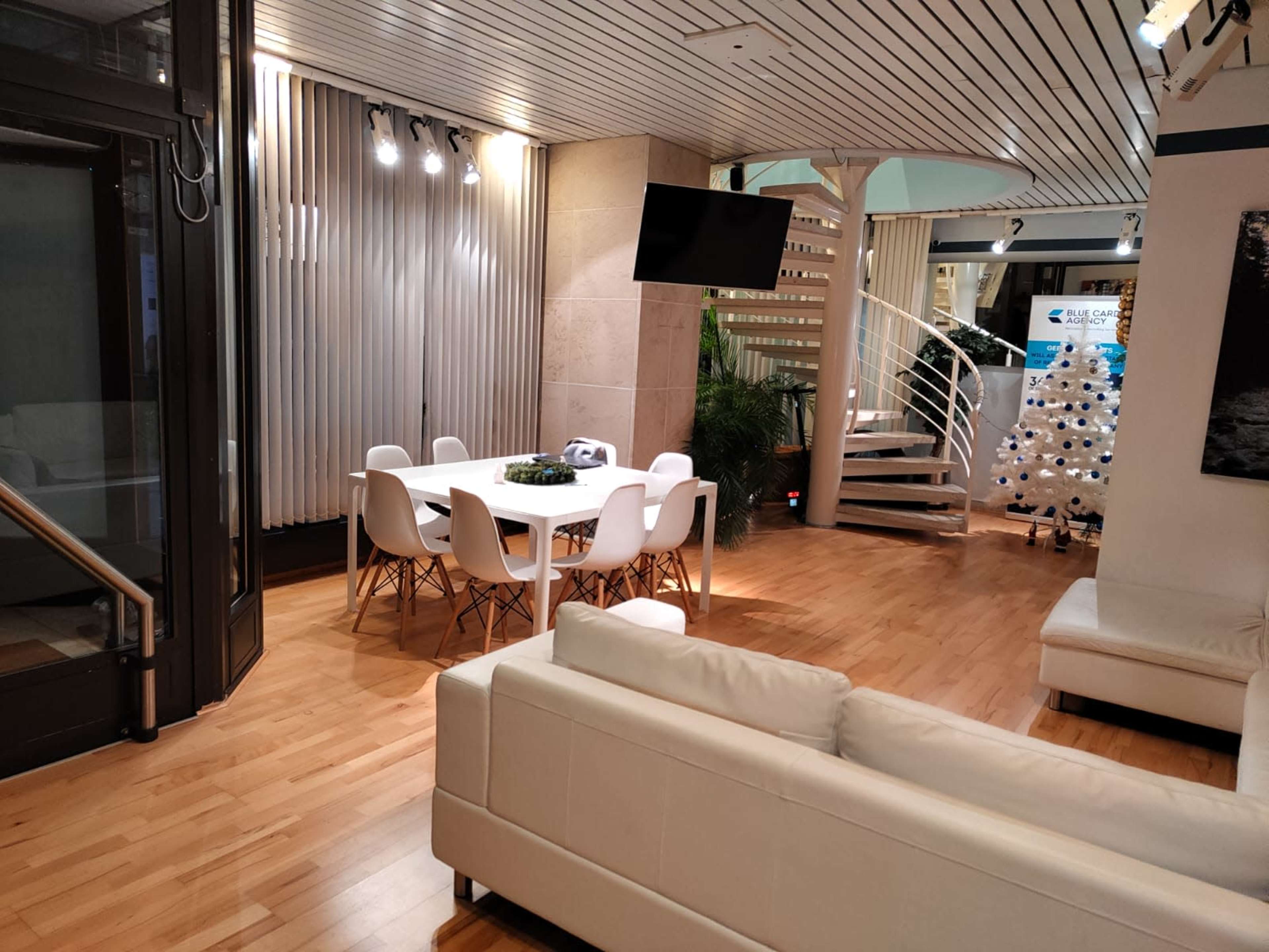 The image shows a modern interior space featuring a dining area with a white table and chairs, a seating area with a white sofa, and a spiral staircase leading to an upper level.