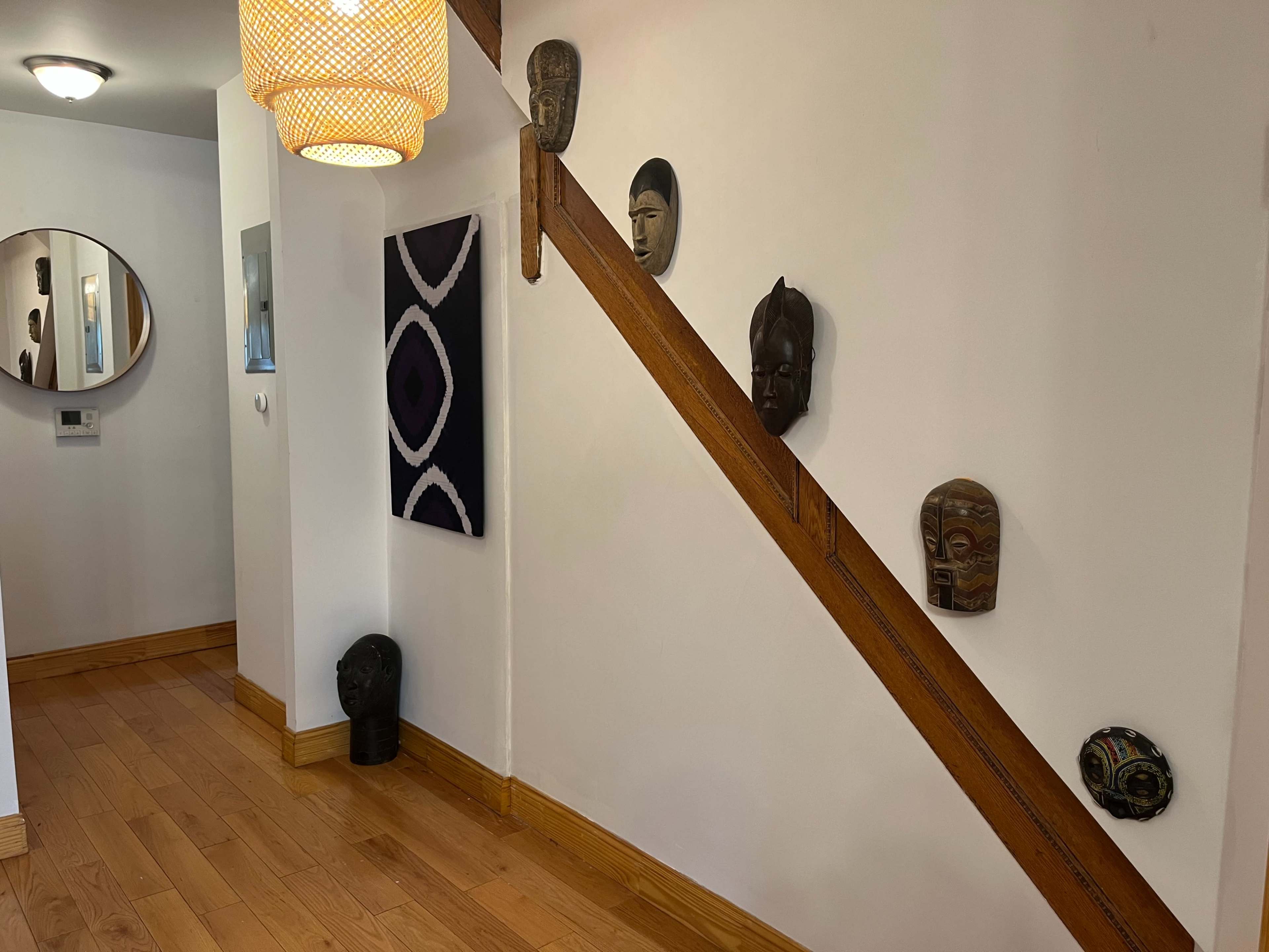 A hallway features wooden flooring, a round mirror, an artwork, and several masks mounted on the wall alongside a wooden staircase.