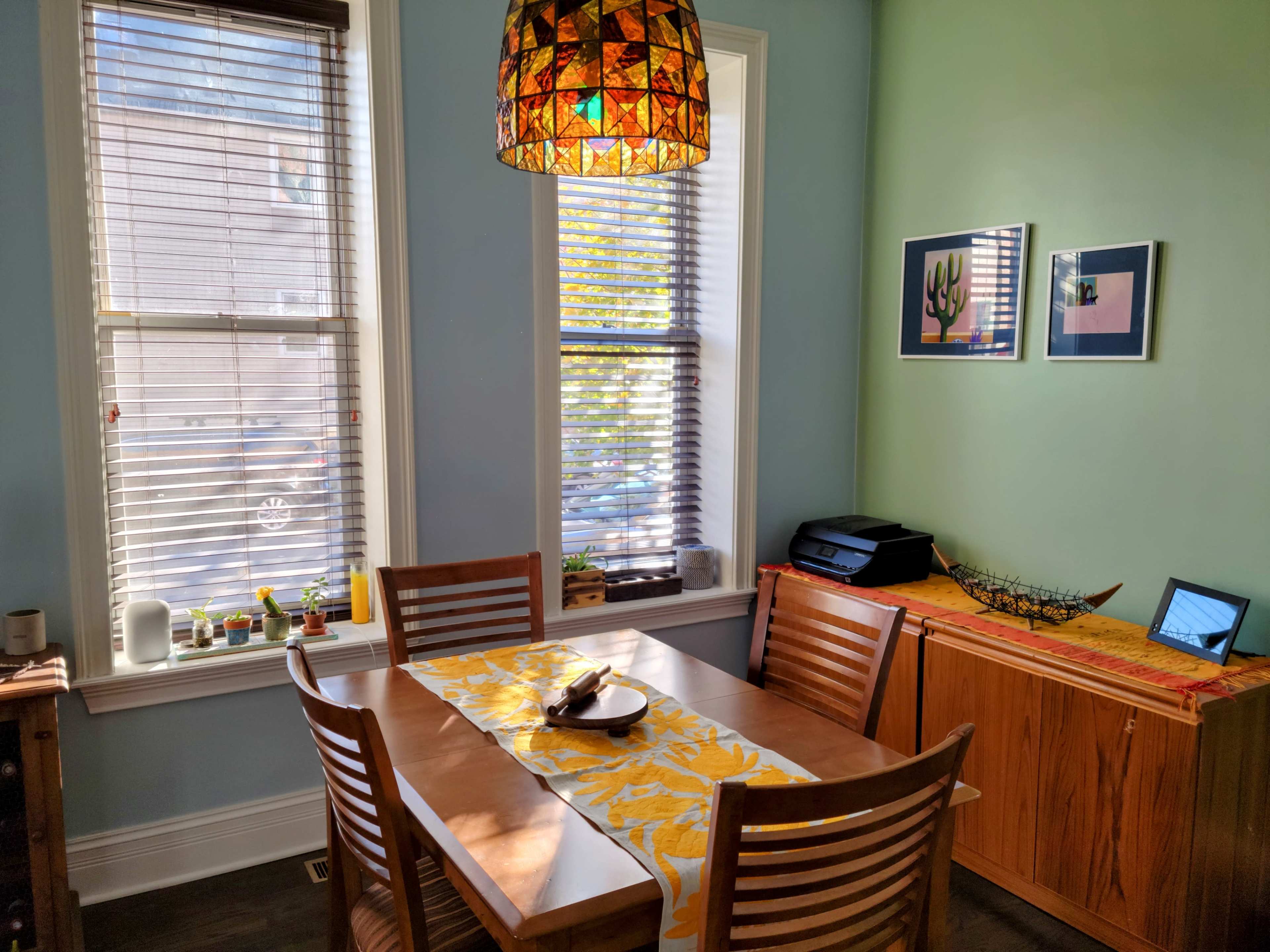 A dining area features a wooden table with a yellow table runner, surrounded by four chairs, and is illuminated by a stained glass pendant light, with sunlight streaming in through two windows.