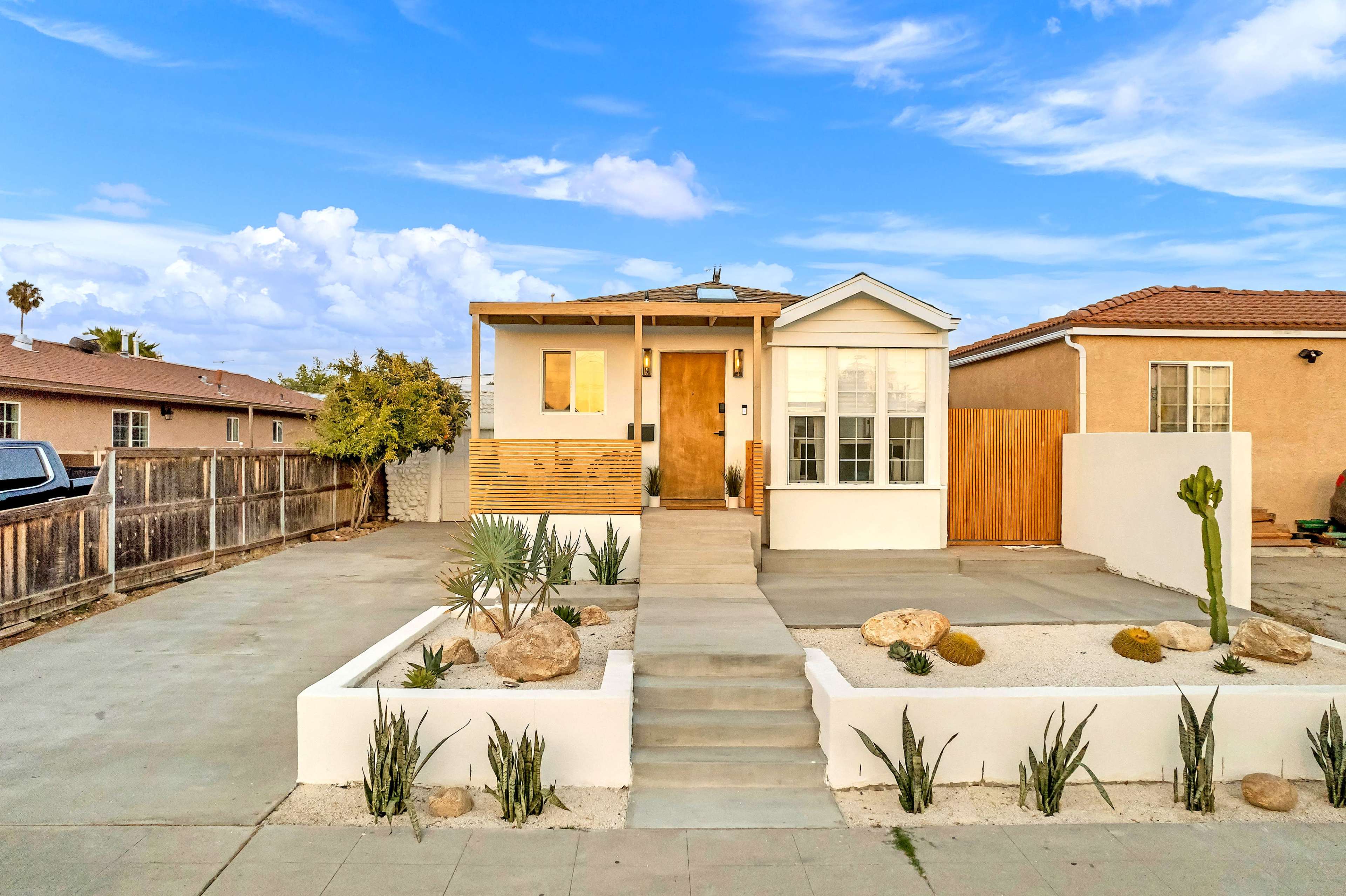 A single-story house with a wooden porch, surrounded by gravel landscaping and desert plants, is set against a blue sky with clouds.