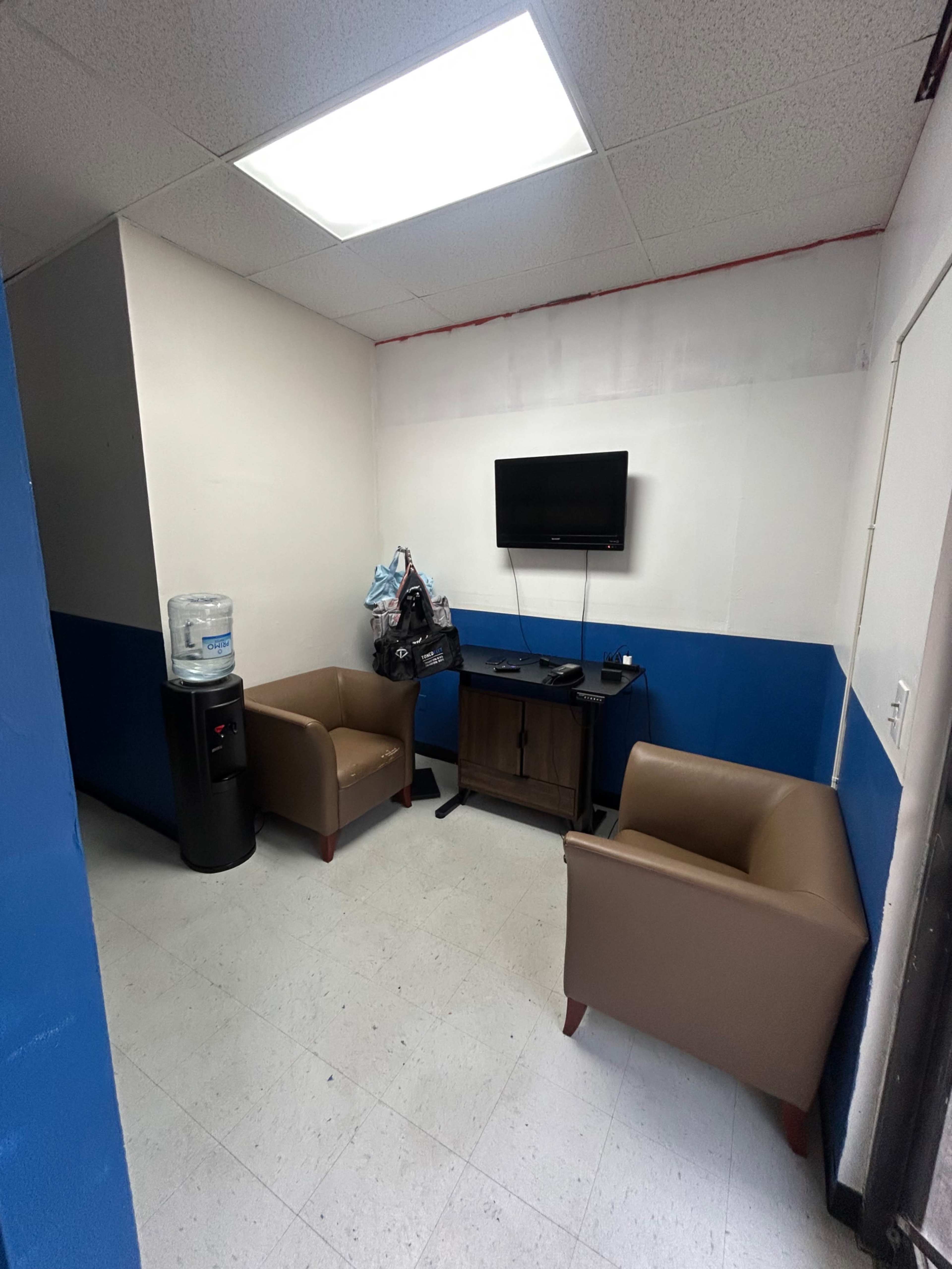 The image shows a small waiting area with two tan chairs, a black television mounted on the wall, a water cooler, and a wooden cabinet.