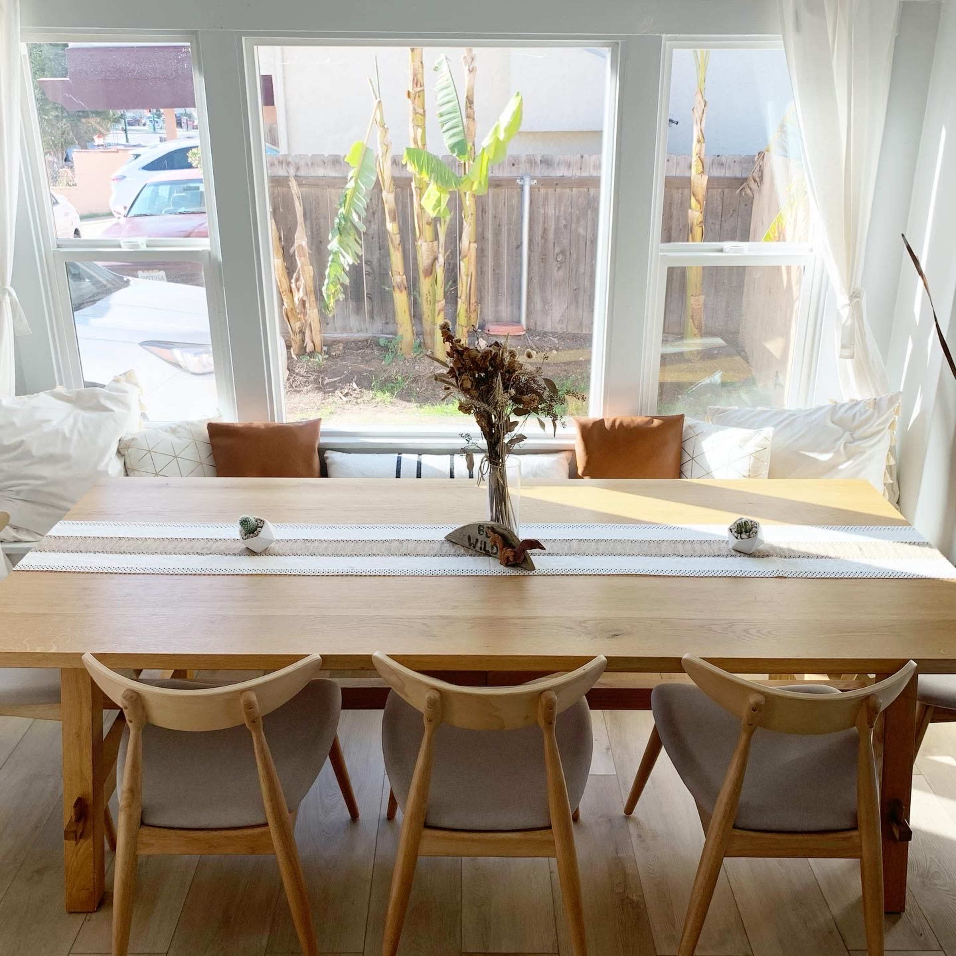 A large wooden dining table is set by a window, surrounded by four chairs and decorated with a runner and a small centerpiece.