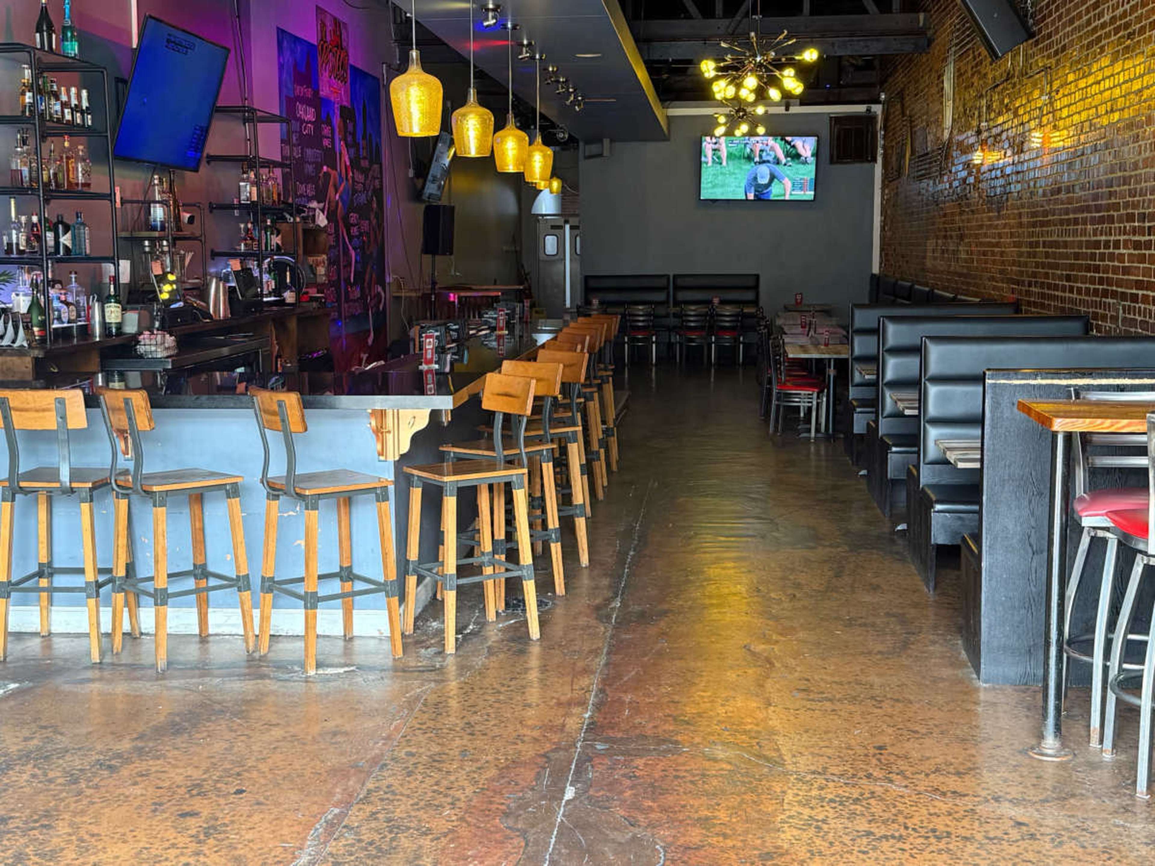 The image shows the interior of a modern bar or restaurant featuring a long bar counter with stools and a seating area along the walls, illuminated by pendant lights and a television screen mounted above.