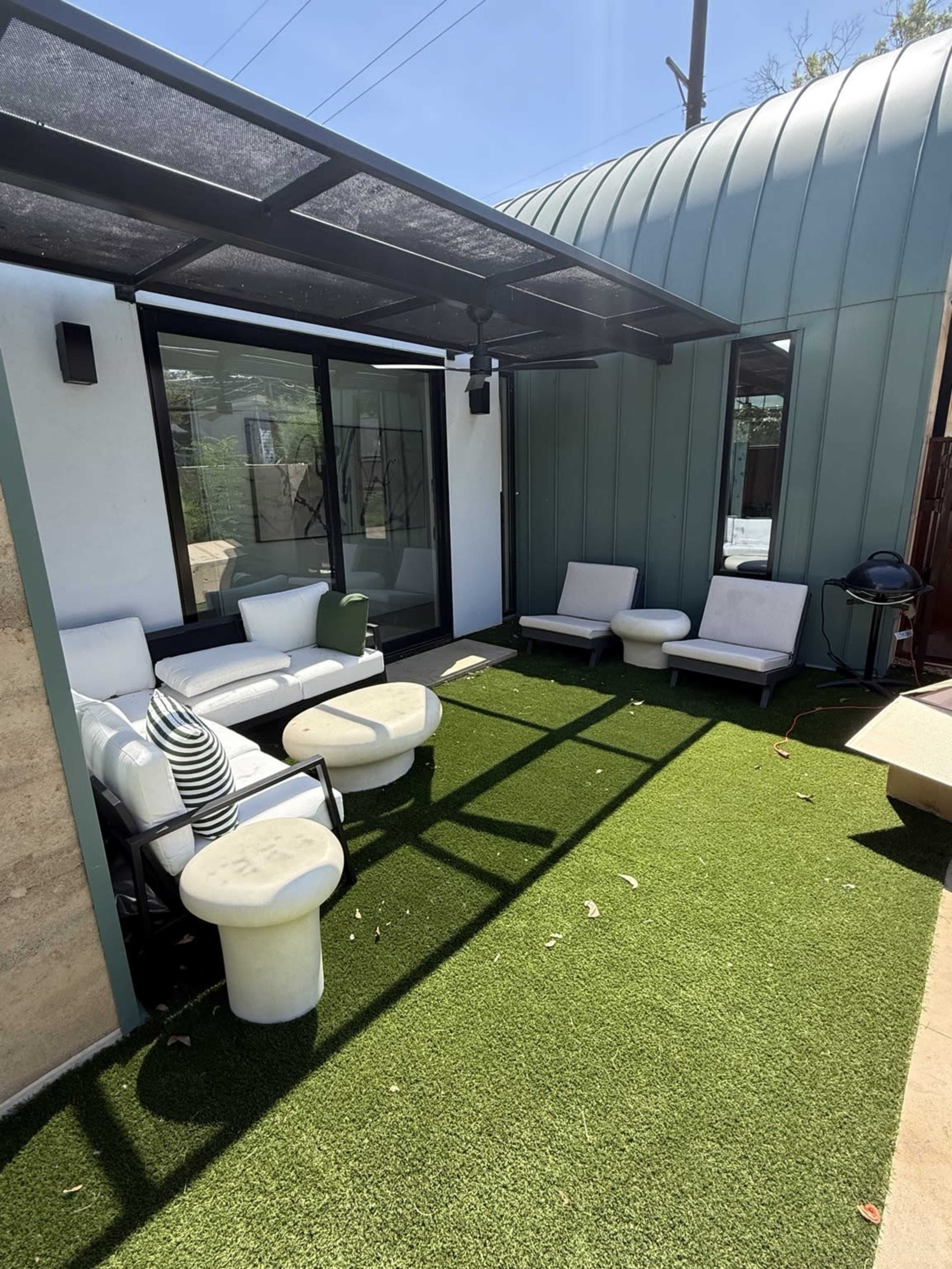 The image shows a modern outdoor patio area with a seating arrangement, tables, and artificial grass underneath a metal awning.