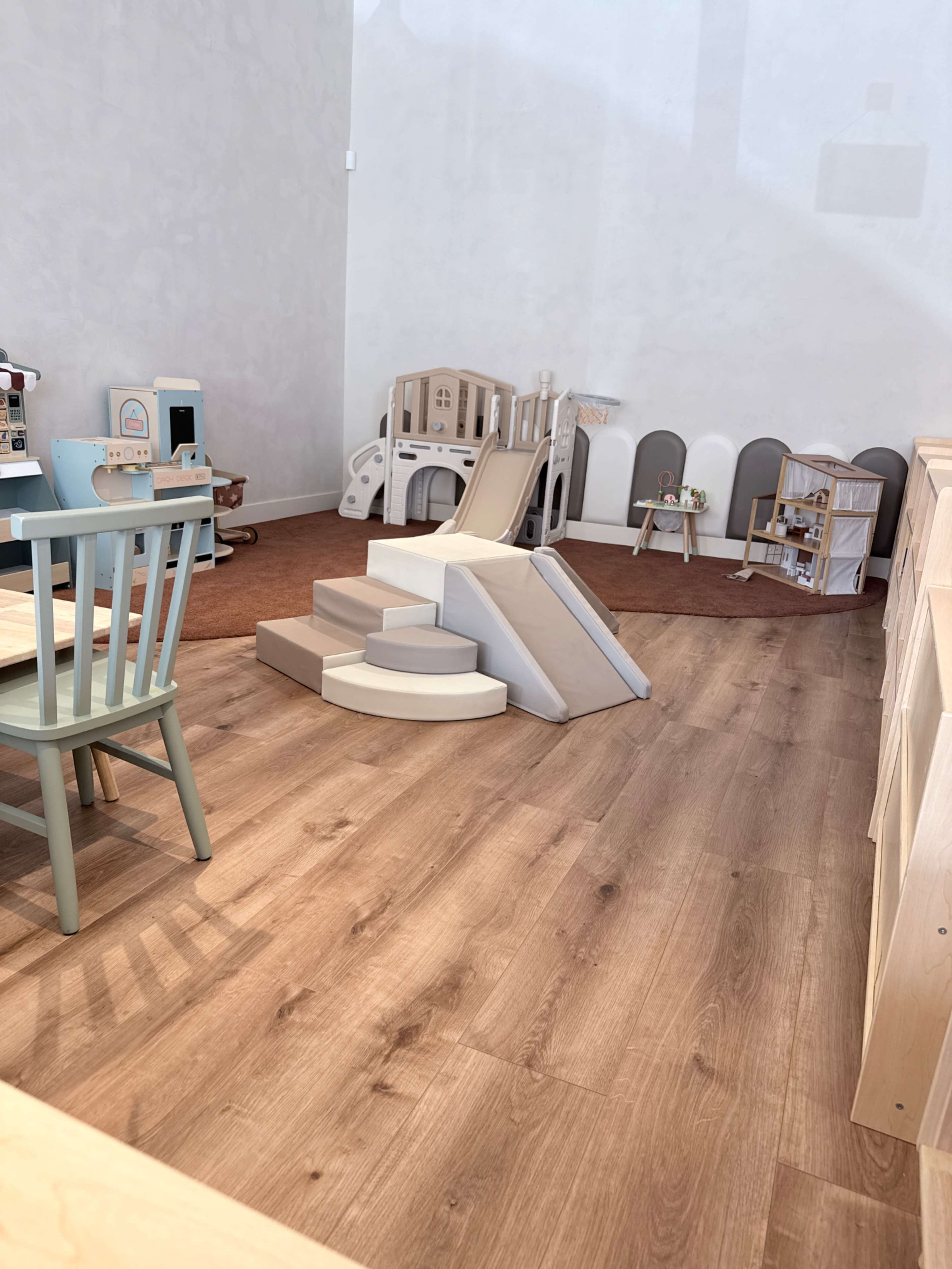 The image shows a playroom with wooden flooring, featuring a small slide and climbing structure, along with various toys and furniture scattered around.