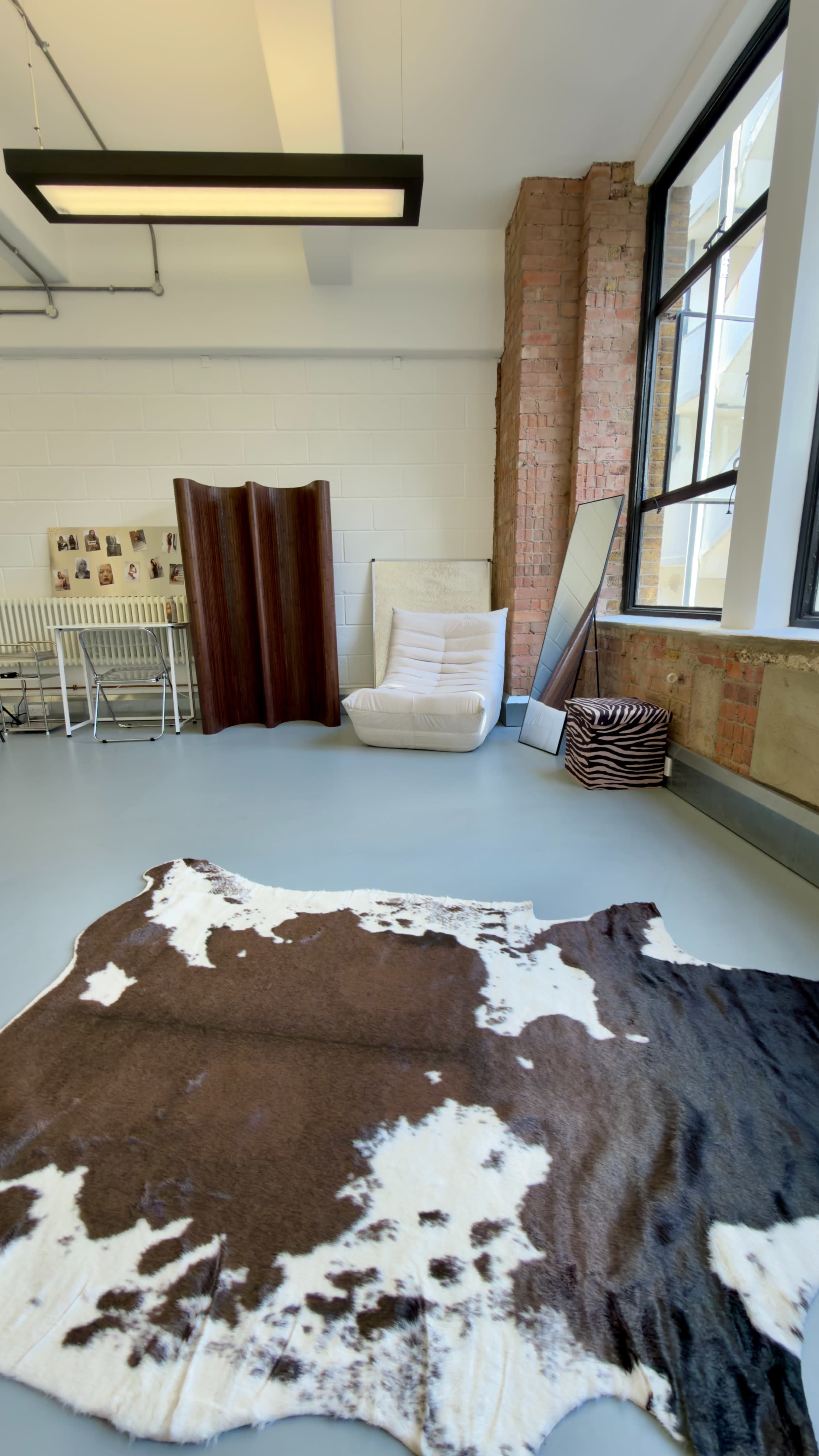 The image shows a spacious interior room with large windows, featuring a brown and white cowhide rug on the floor, a white chair, a mirror, and a folding screen along with various other furnishings and decor elements.