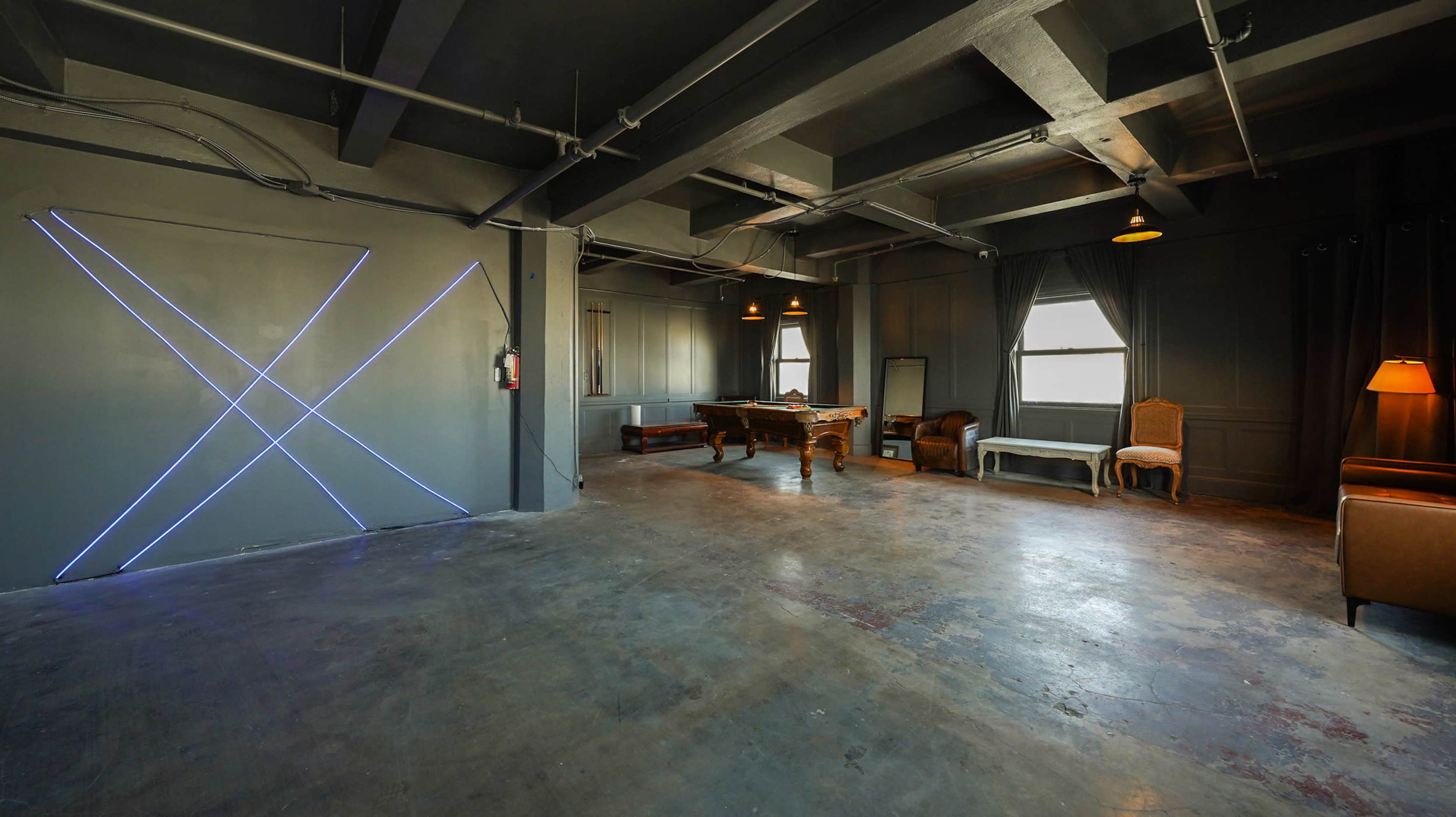 A spacious, empty room with gray walls, a polished concrete floor, and a few pieces of furniture, including a billiards table and chairs, illuminated by wall-mounted lights.