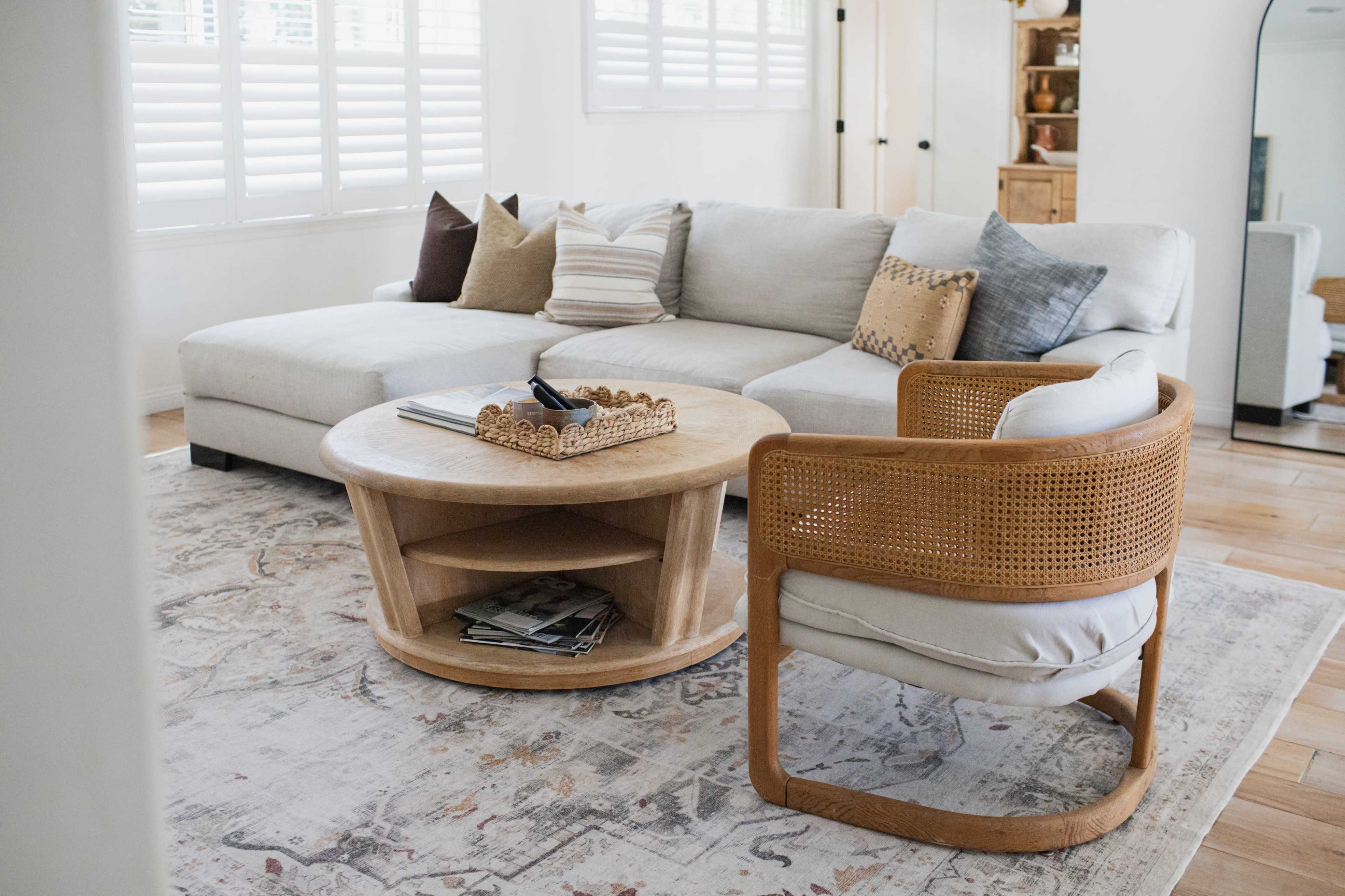 The image shows a living room featuring a light-colored sectional sofa, a round coffee table with decorative items, and a wooden chair set on a patterned rug.