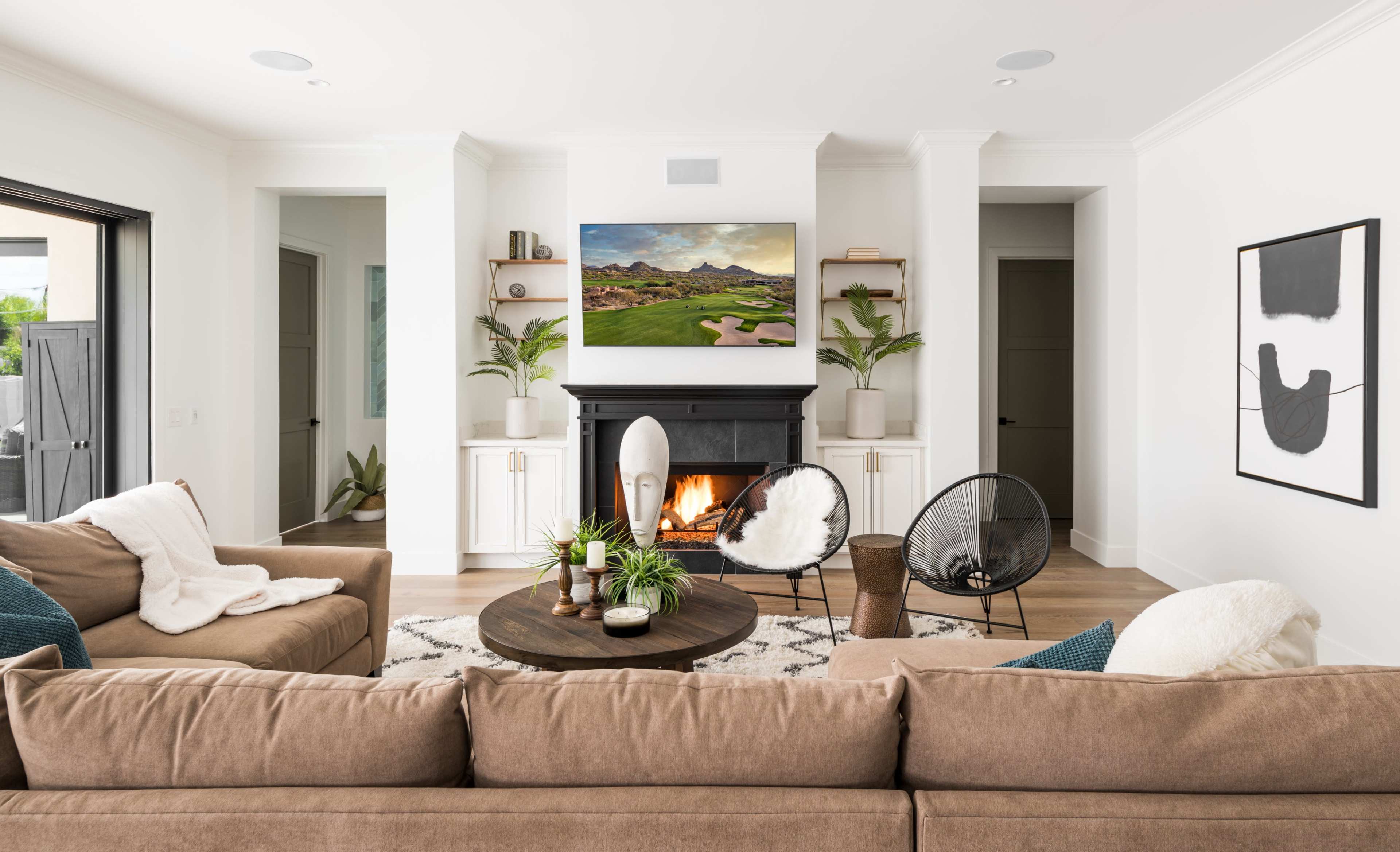 A modern living room features a central coffee table, comfortable seating, and a fireplace framed by a large painting and decorative plants.