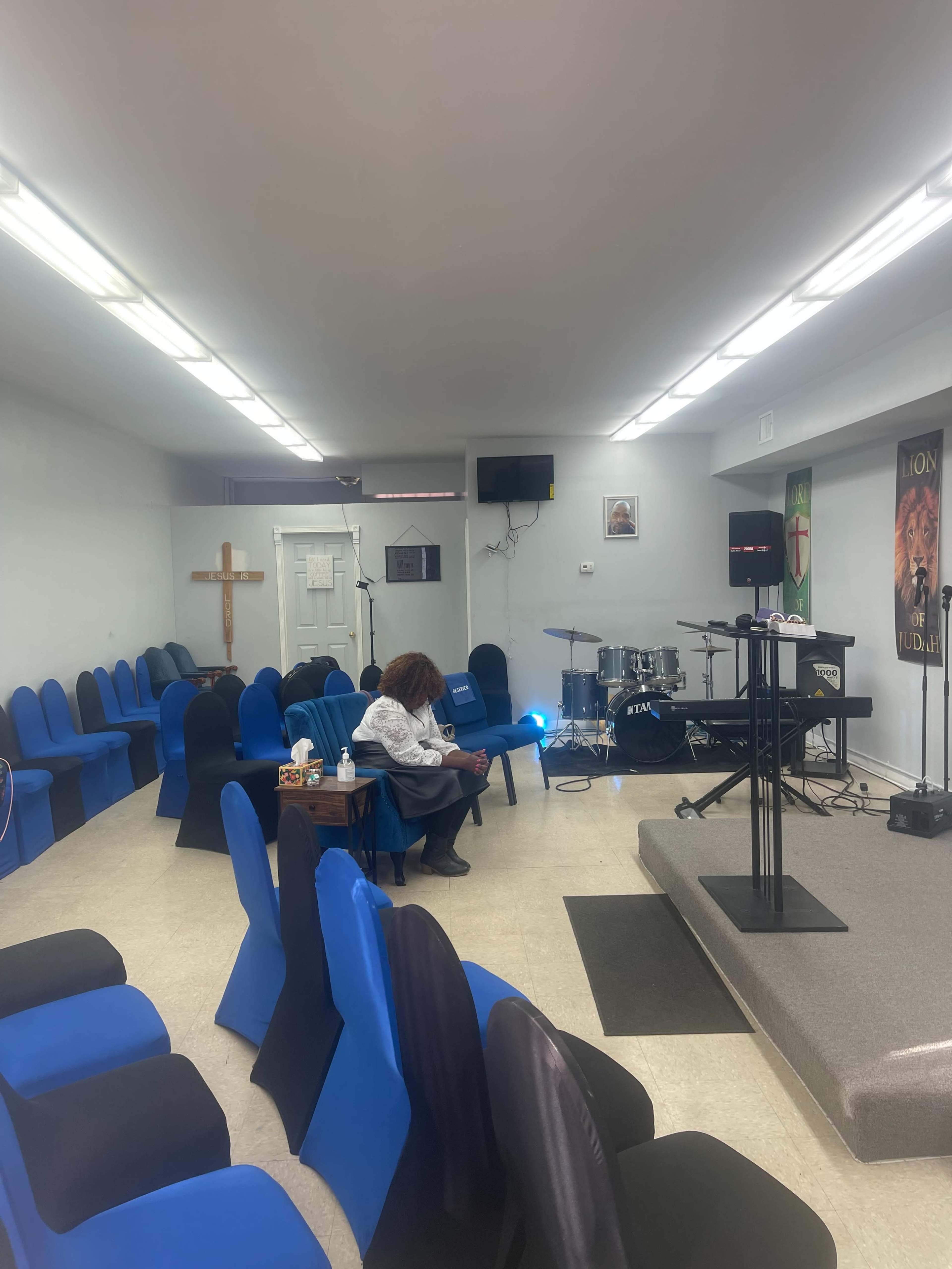 A person is seated in a row of blue chairs inside a small, sparsely decorated church, with a podium and musical instruments in the background.
