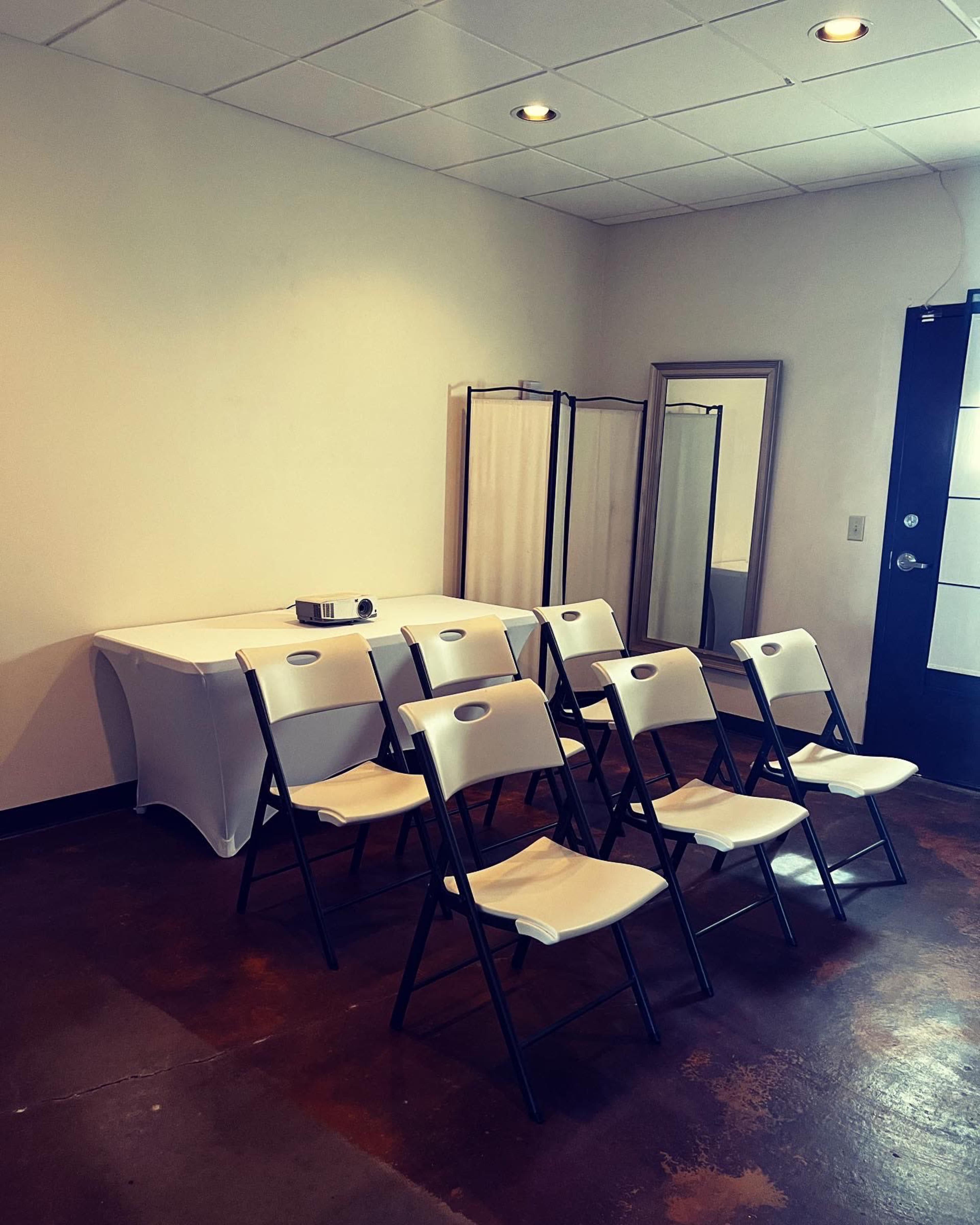 A small meeting room features a row of folding chairs facing a table with a projector and a mirror on the side.