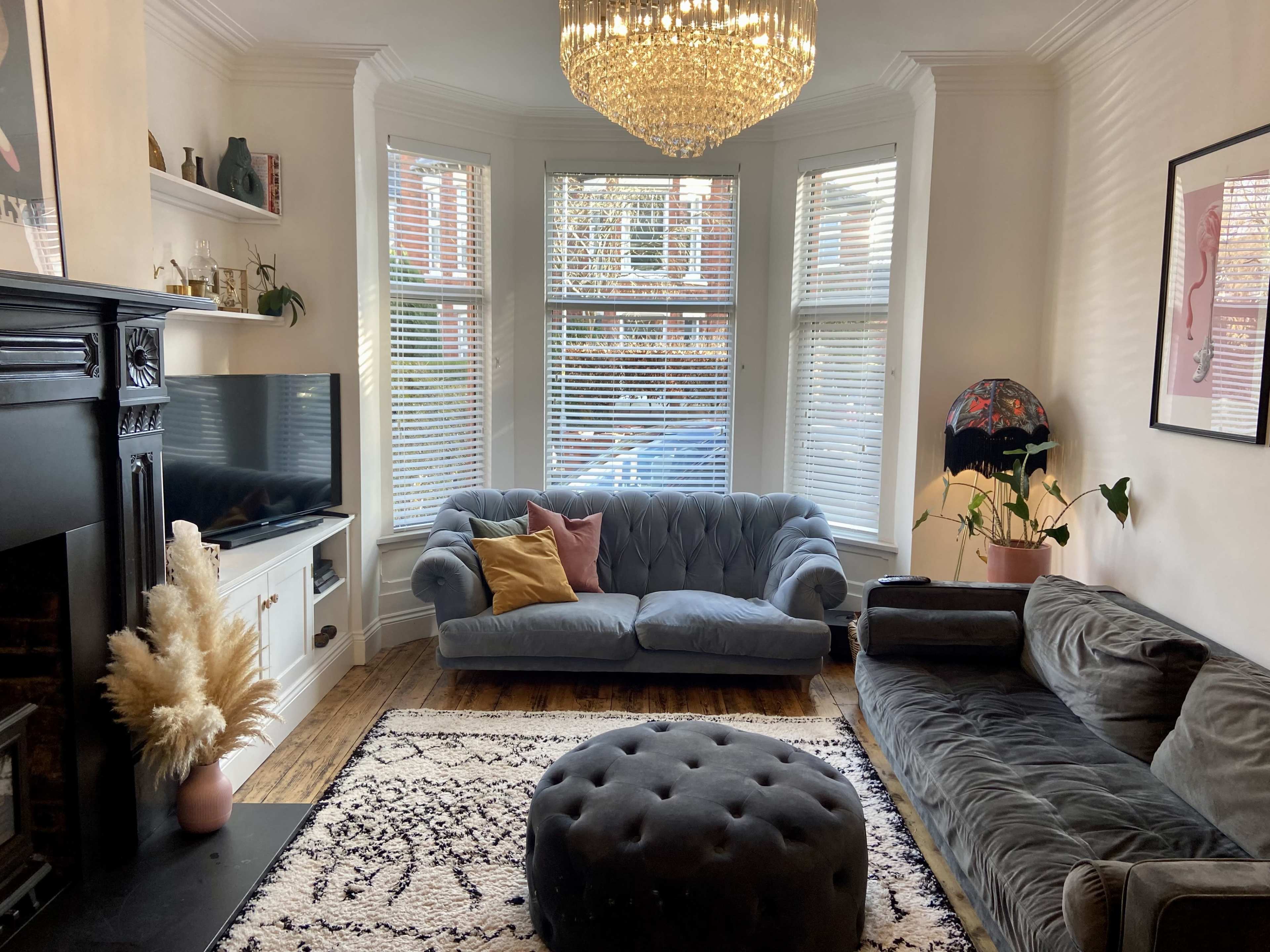 The room features a stylish seating area with a tufted blue sofa and a large round ottoman, illuminated by a chandelier and natural light from bay windows.