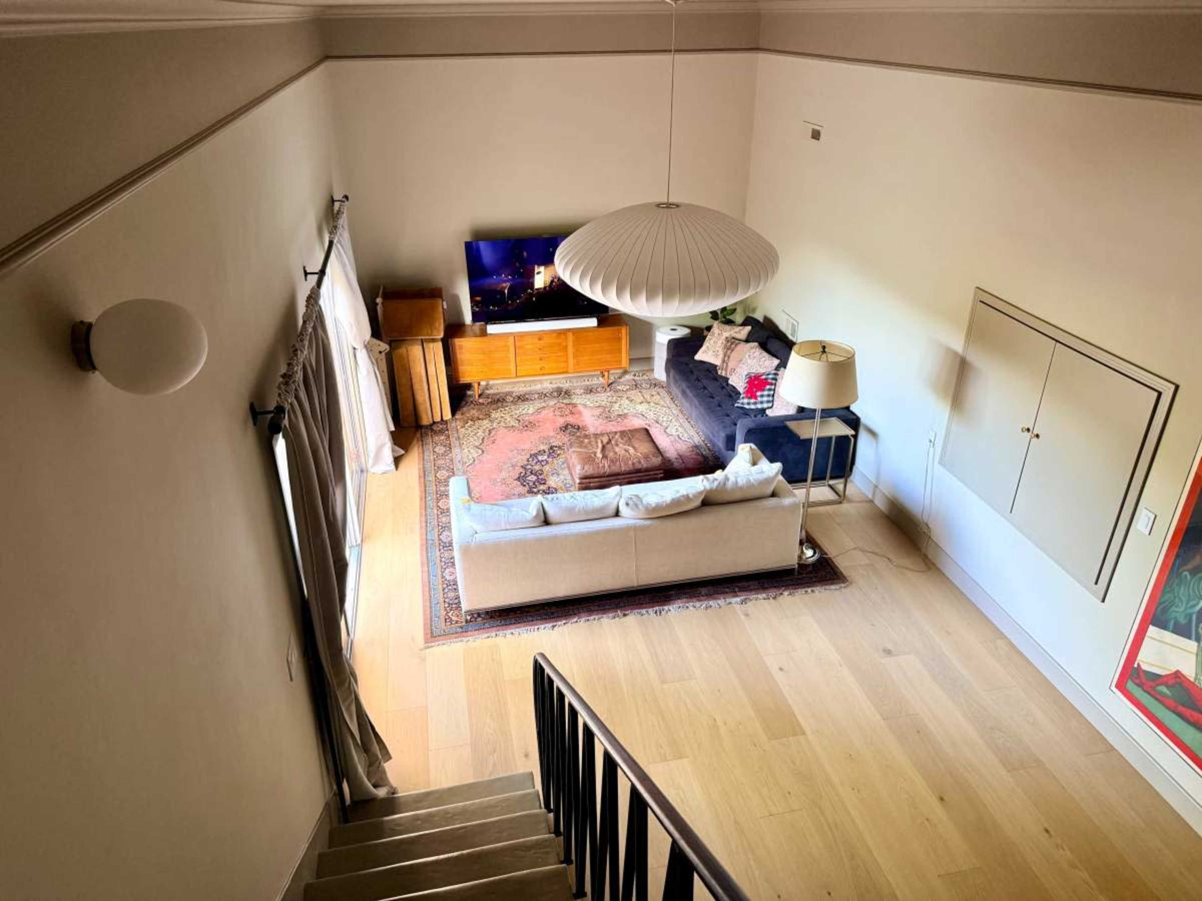 The image shows a living room viewed from above, featuring a couch, a TV mounted on the wall, a lamp, and a wooden cabinet on a patterned rug.