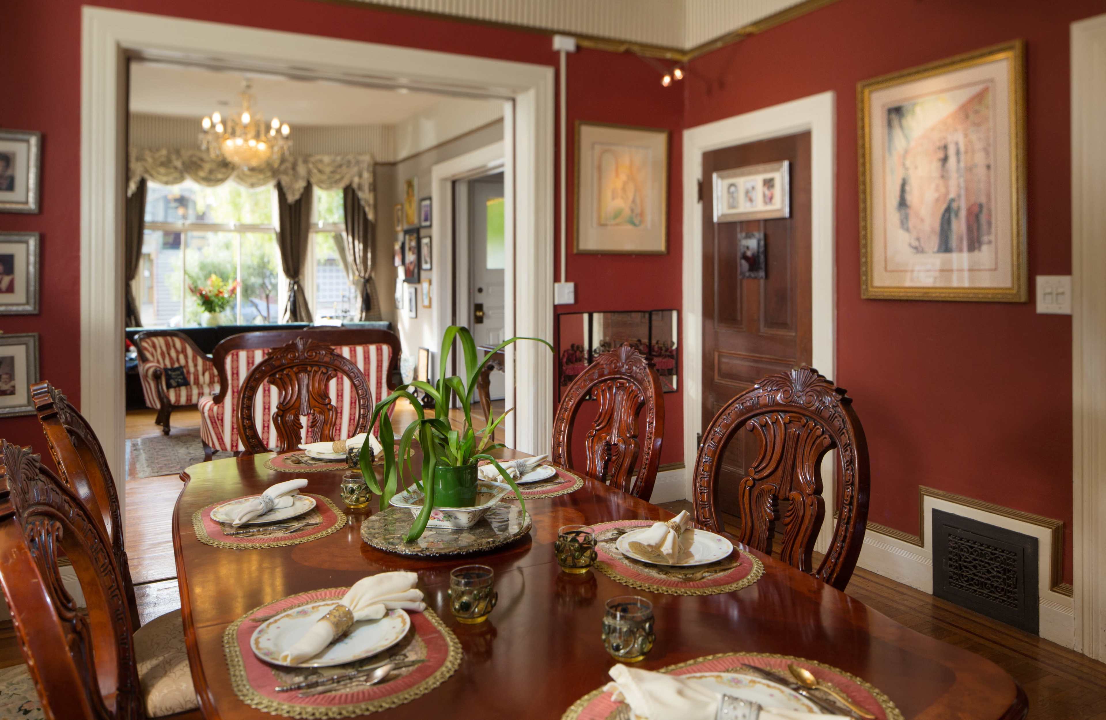 A dining room with a large wooden table set for a meal, surrounded by intricately carved wooden chairs and decorated with framed artworks on the walls.