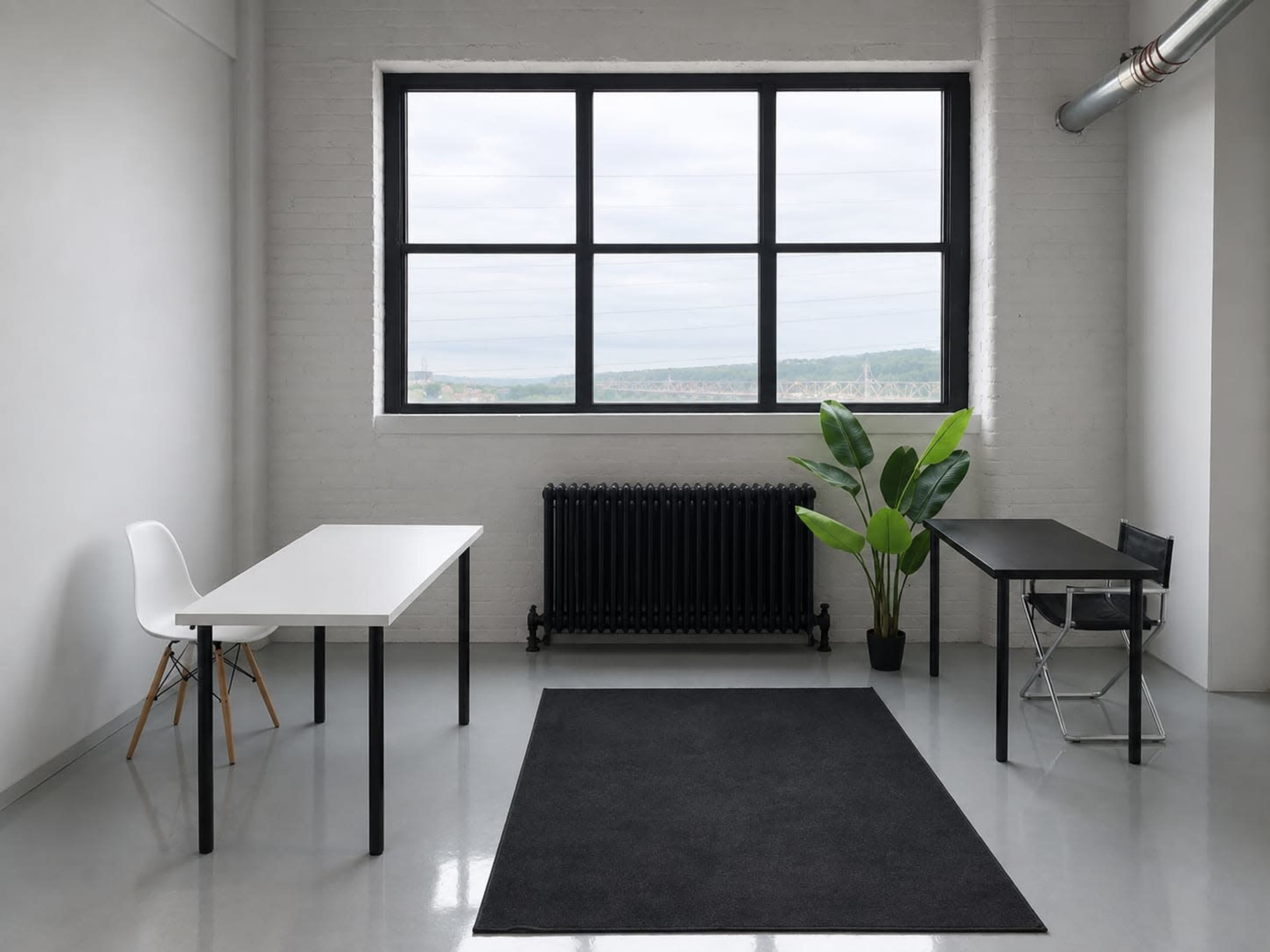 The image shows a minimalistic office space featuring two desks, a radiator, a potted plant, a black rug, and a large window with a view.