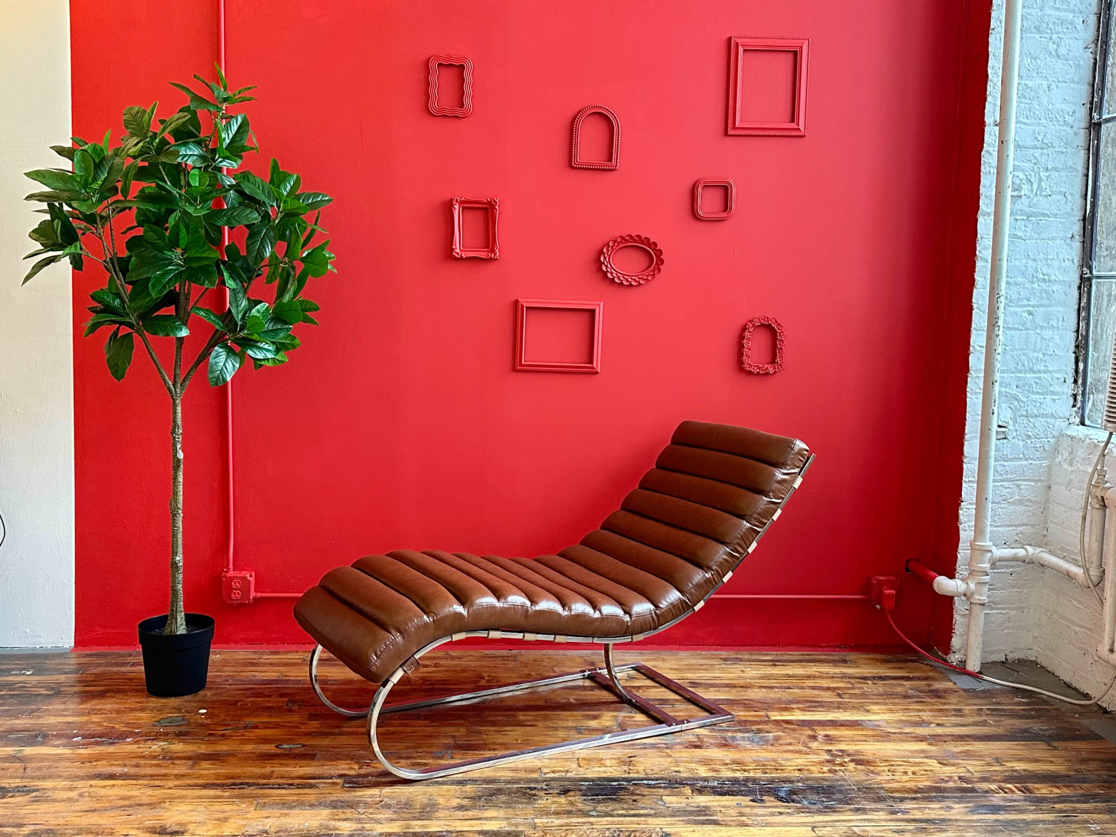 A brown chaise lounge is positioned against a bright red wall adorned with various empty picture frames, alongside a potted green plant.
