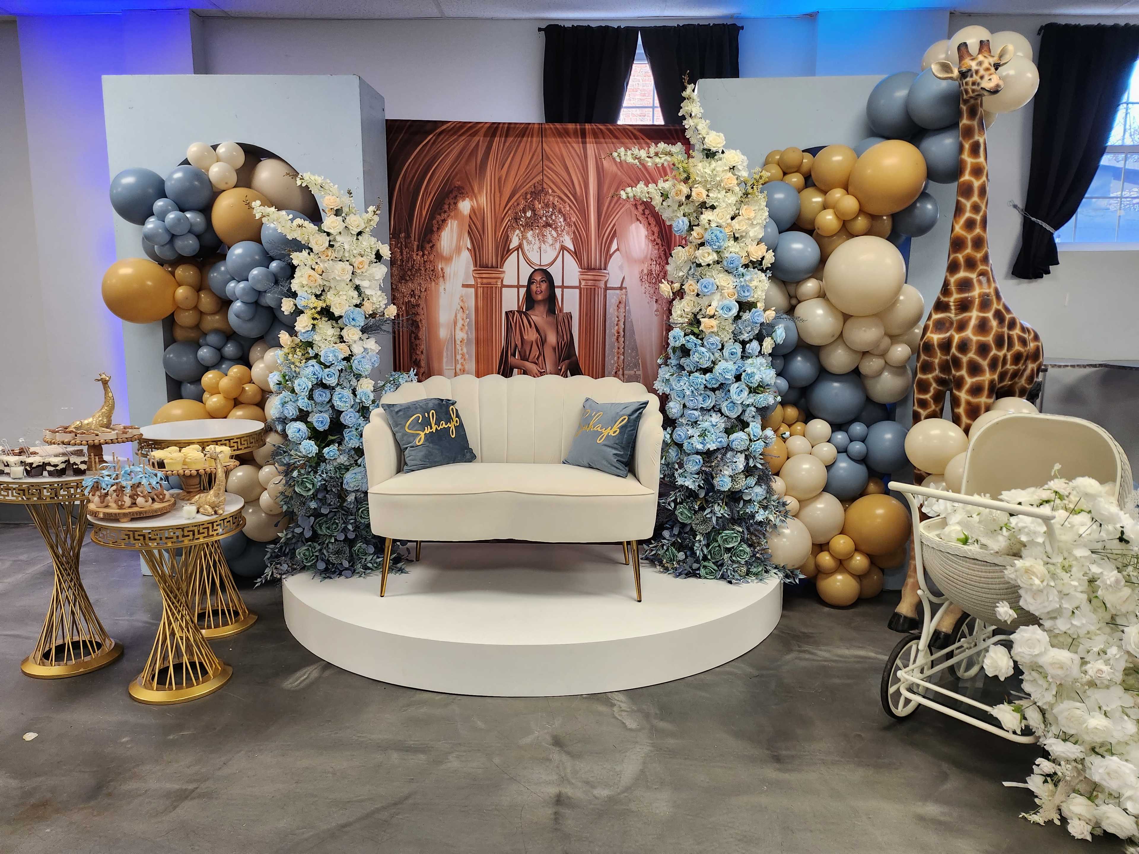The scene features an ornate backdrop with a seated area adorned with balloons, floral arrangements, and a decorative giraffe, set in a spacious hall.