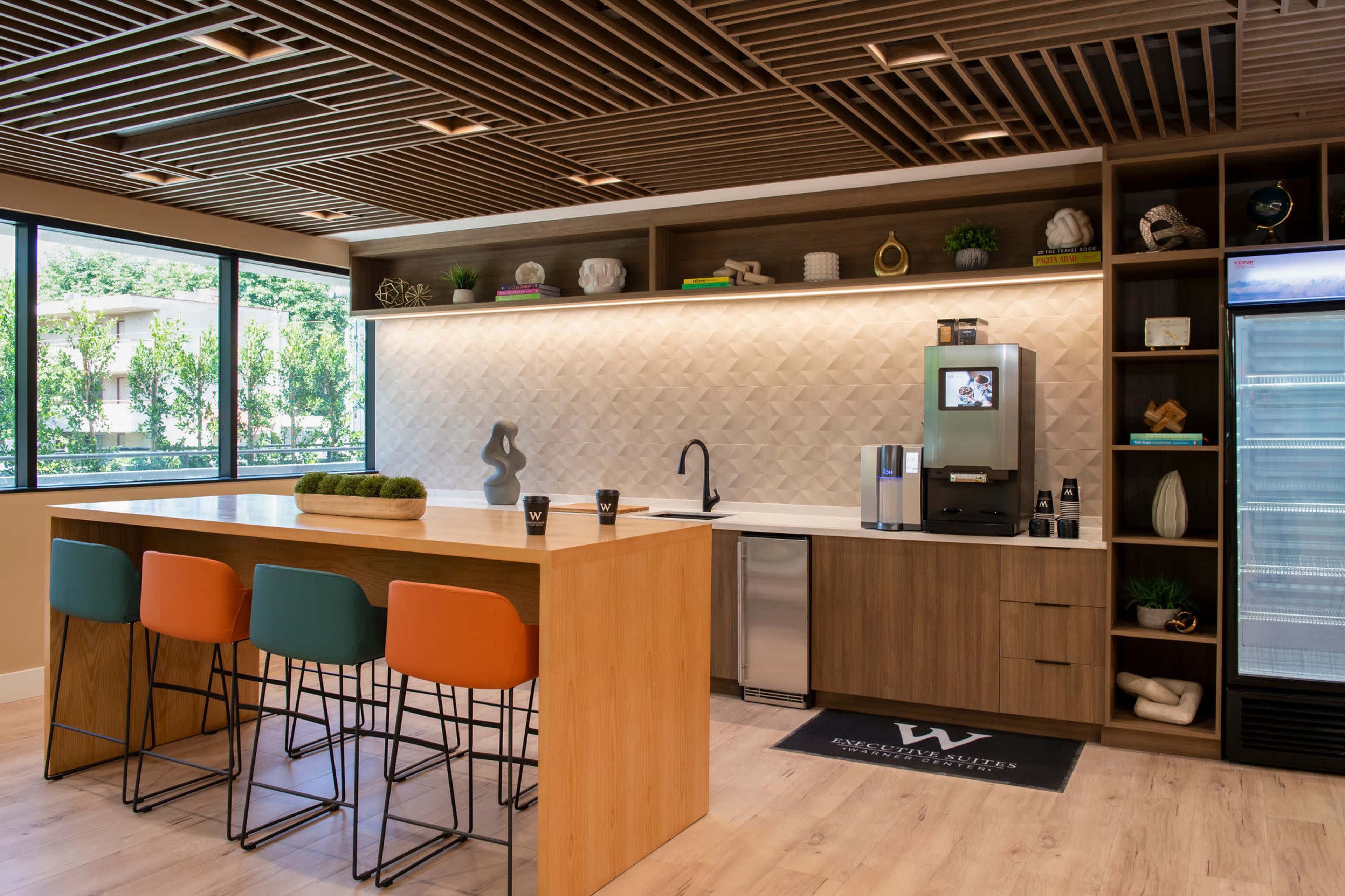 The image shows a modern kitchen area with a wooden island, colorful bar stools, and a coffee machine against a textured wall with various decorative items.